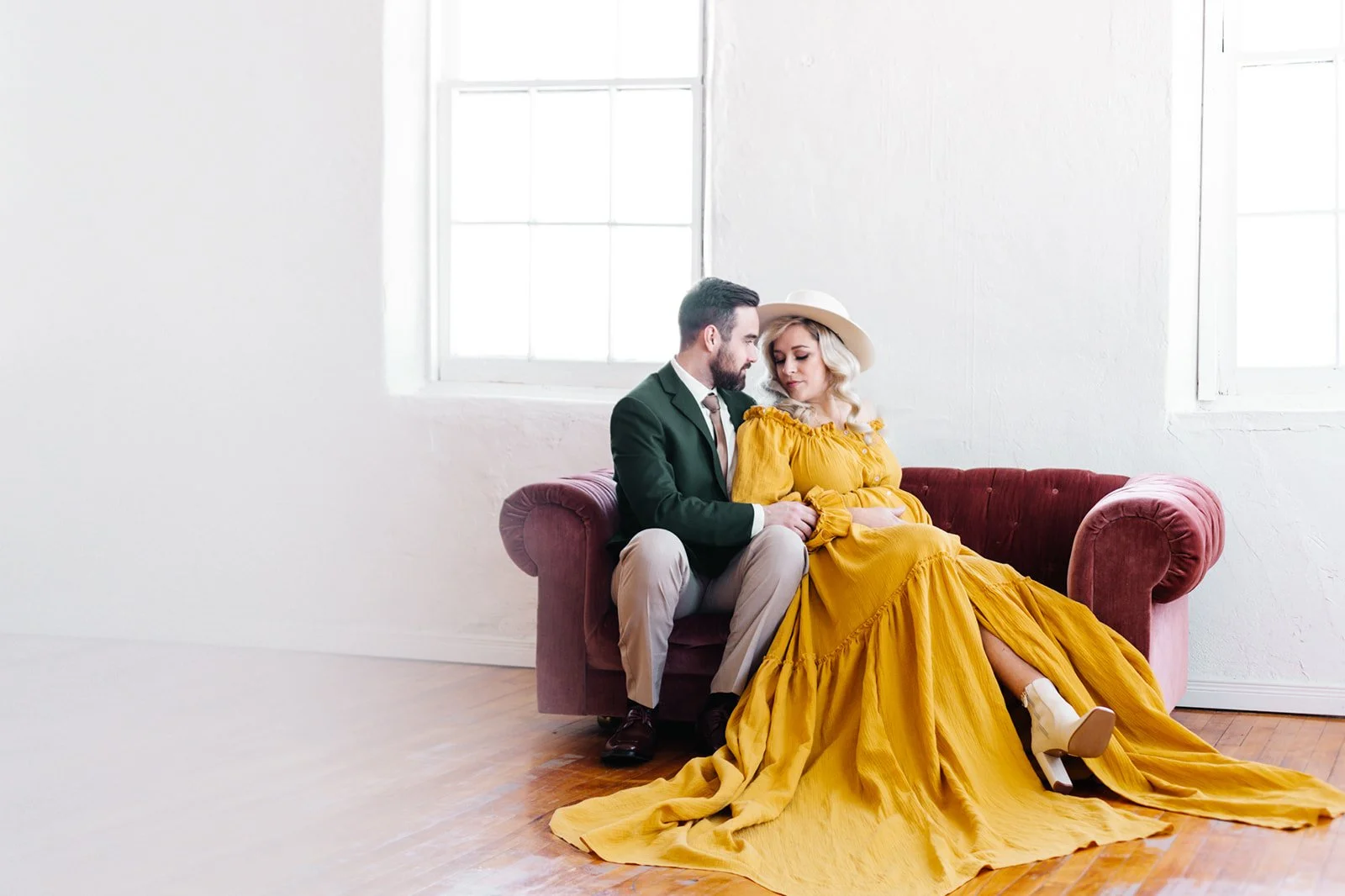 Couple sitting on a velvet sofa in a minimalist room. The woman is wearing a long yellow dress and a hat, while the man is in a green suit with beige pants. They are sitting close together near large windows.