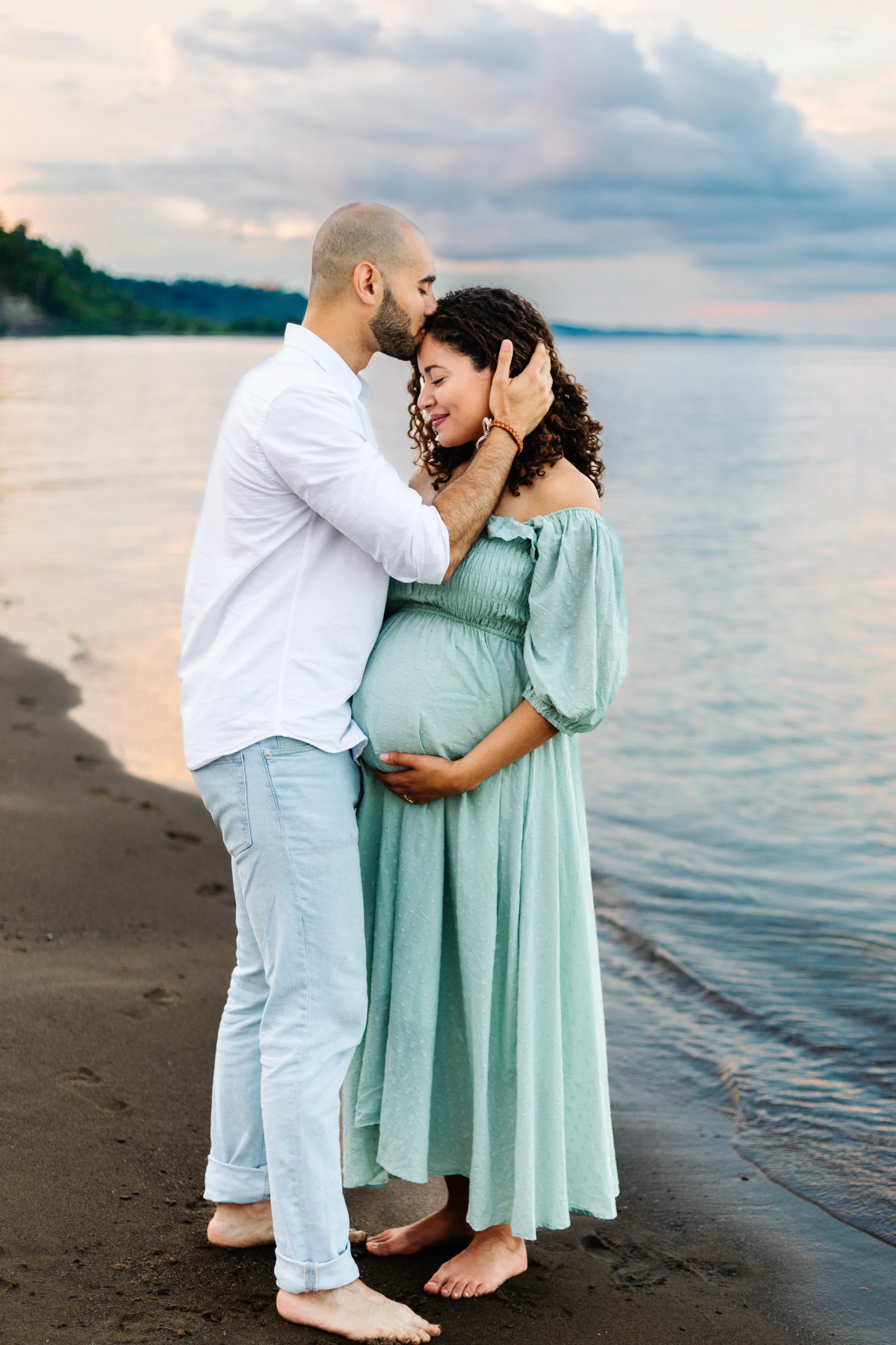 Pregnant couple embracing on the beach, woman wearing a light green dress, man in white shirt and jeans, ocean and sunset in the background.