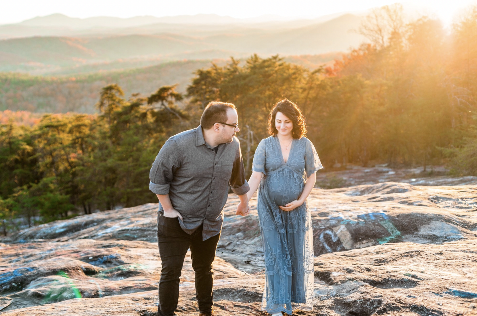 Pregnant woman and man walking on rocky terrain during sunset, holding hands, with forest and mountains in the background.