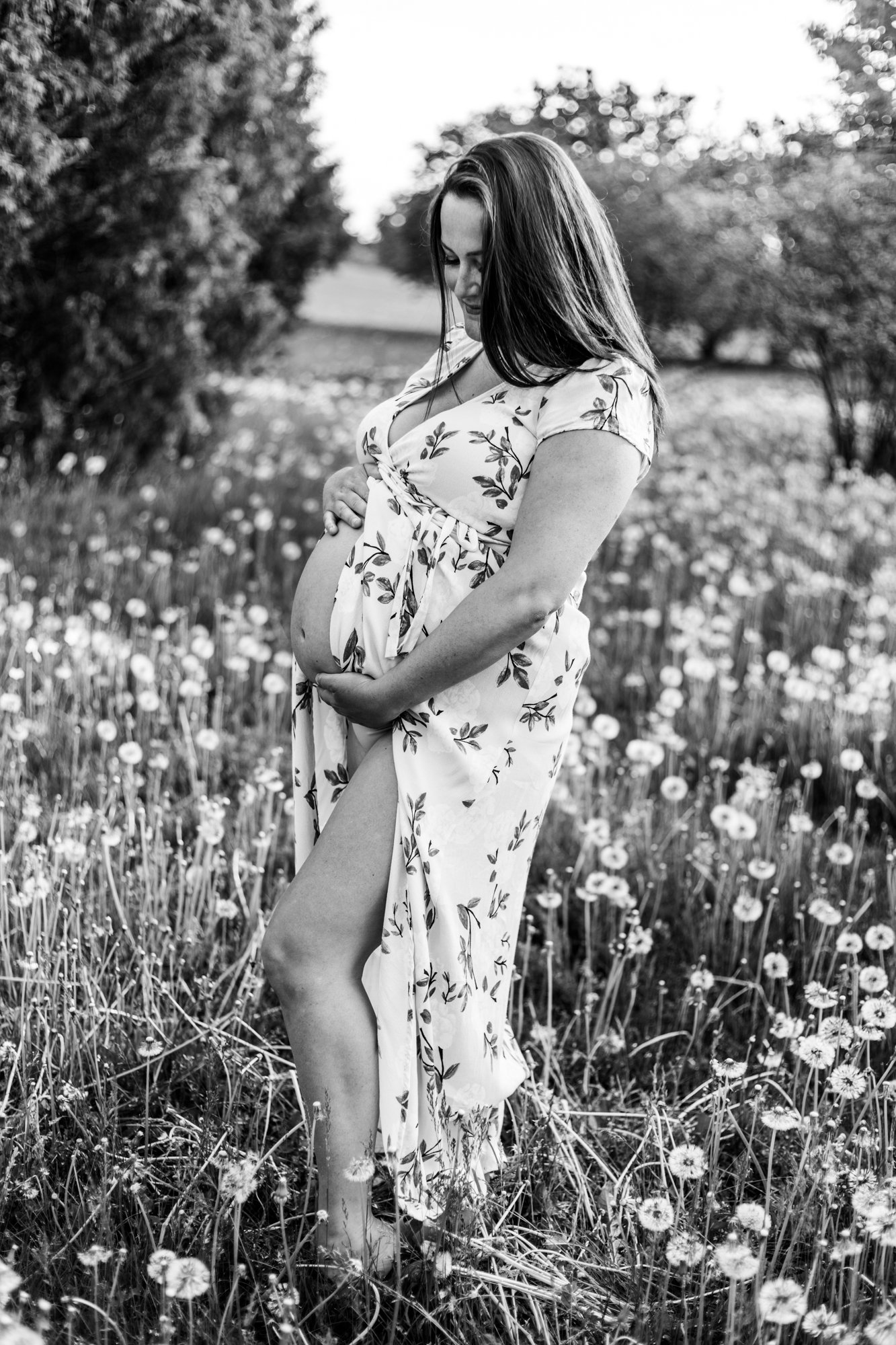 Black and white photo of a pregnant woman in a floral dress standing in a field with flowers