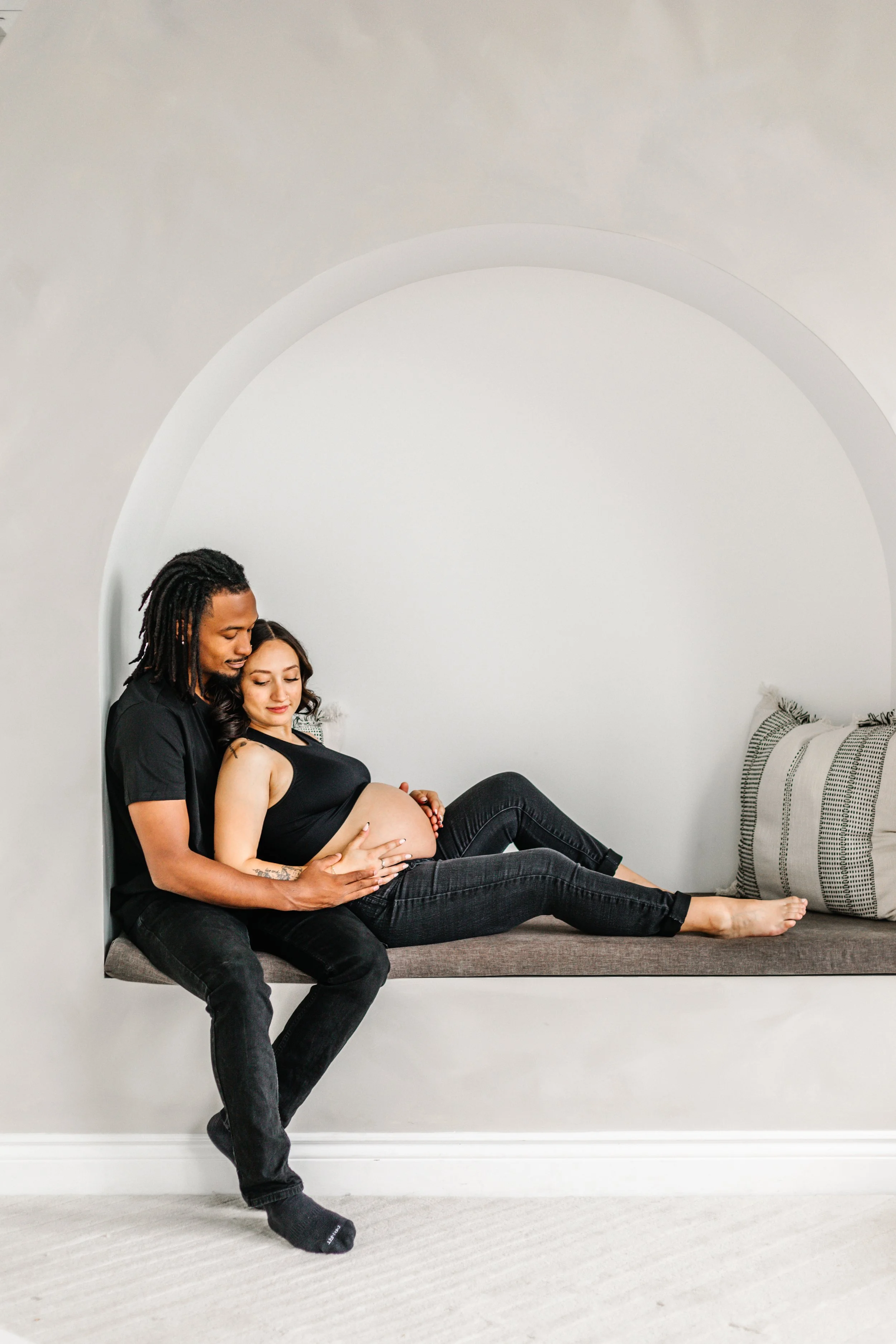 A couple sitting together, with the man embracing the pregnant woman and touching her belly tenderly. They are on a cushioned seat with neutral decor.