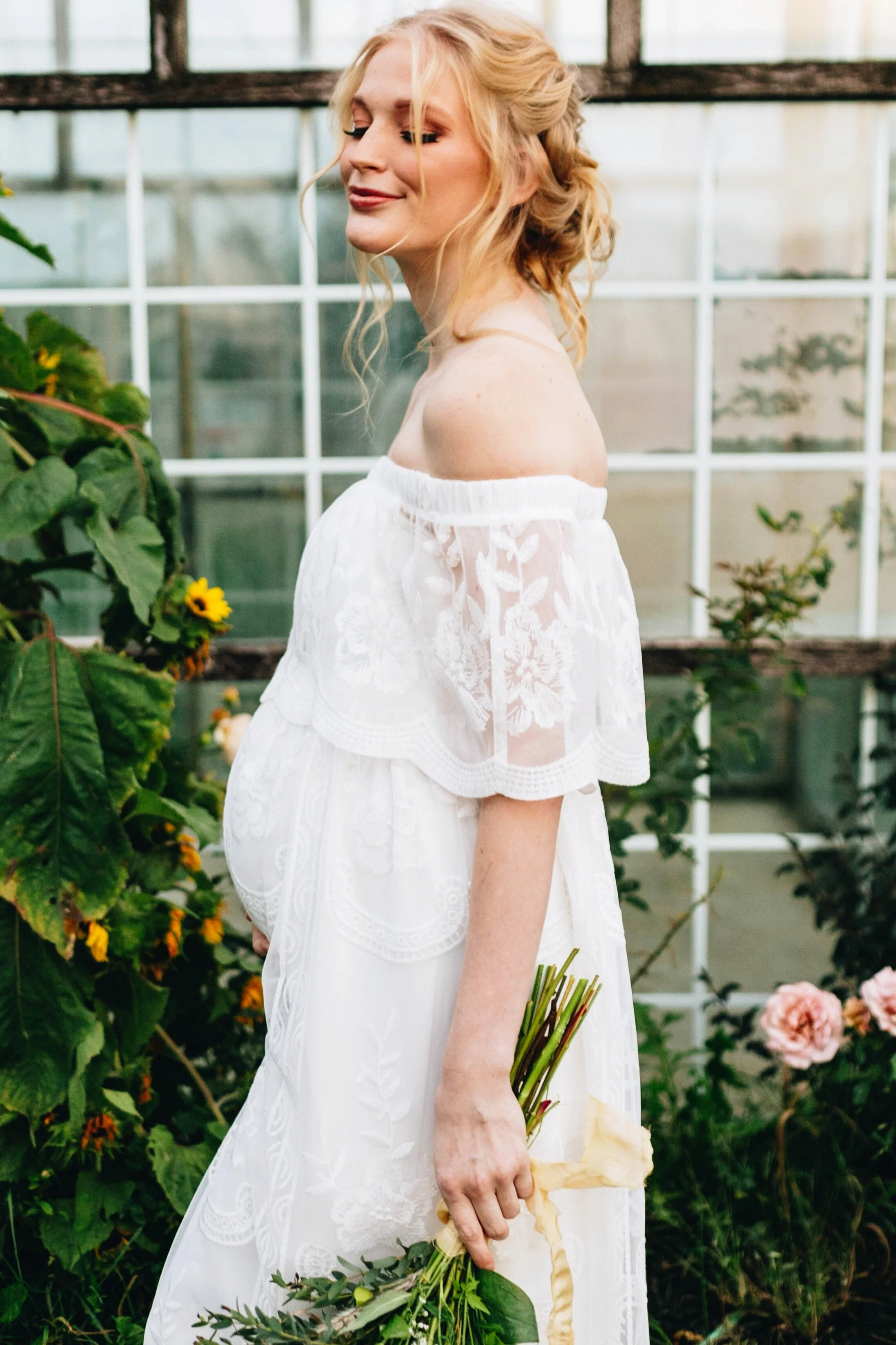 Pregnant woman in white lace dress holding flowers, standing in garden with glass windows and plants.