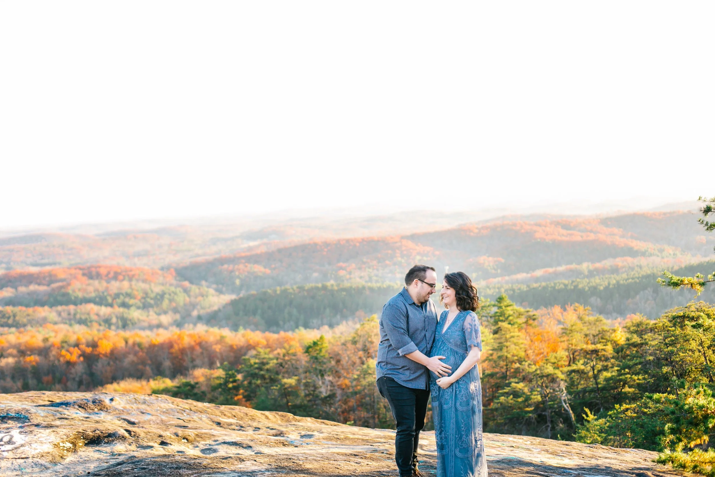 Couple standing on a rocky cliff with a scenic view of autumn foliage in the background, during daytime.