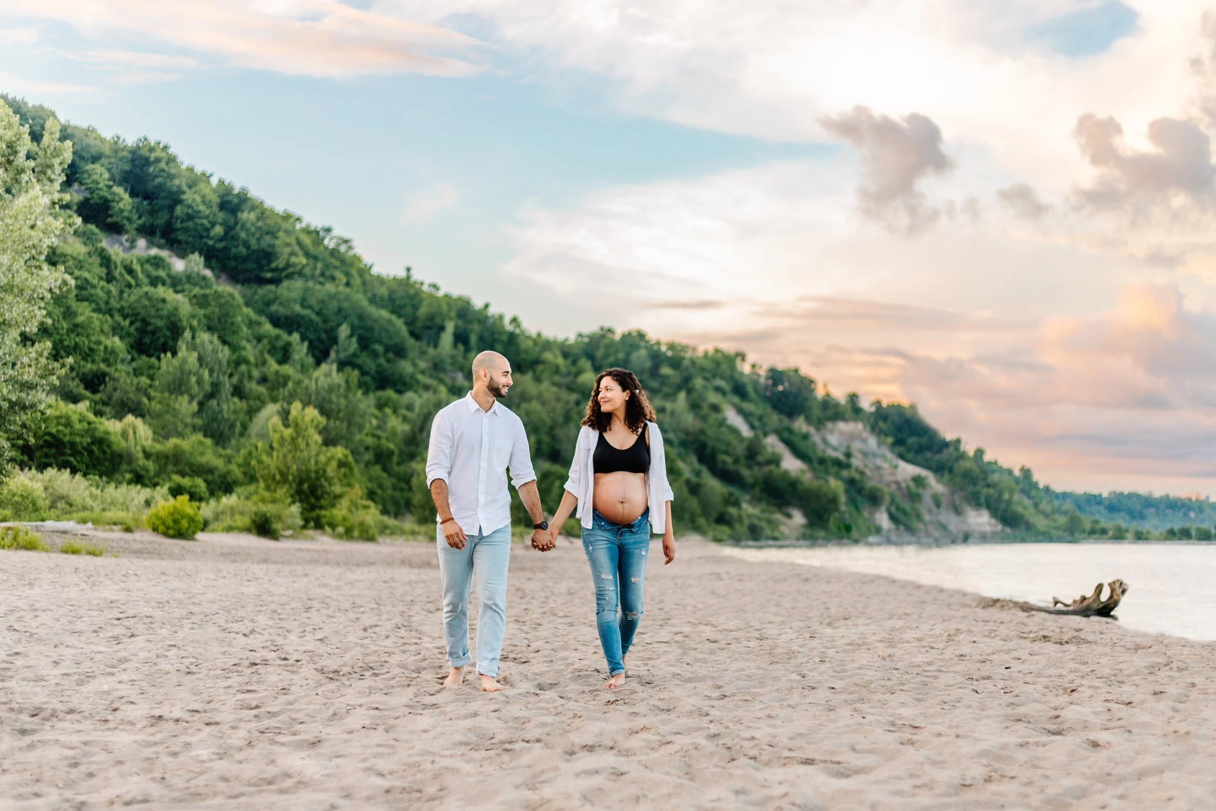 Pregnant couple holding hands on a beach with a scenic backdrop of greenery and a colorful sky.