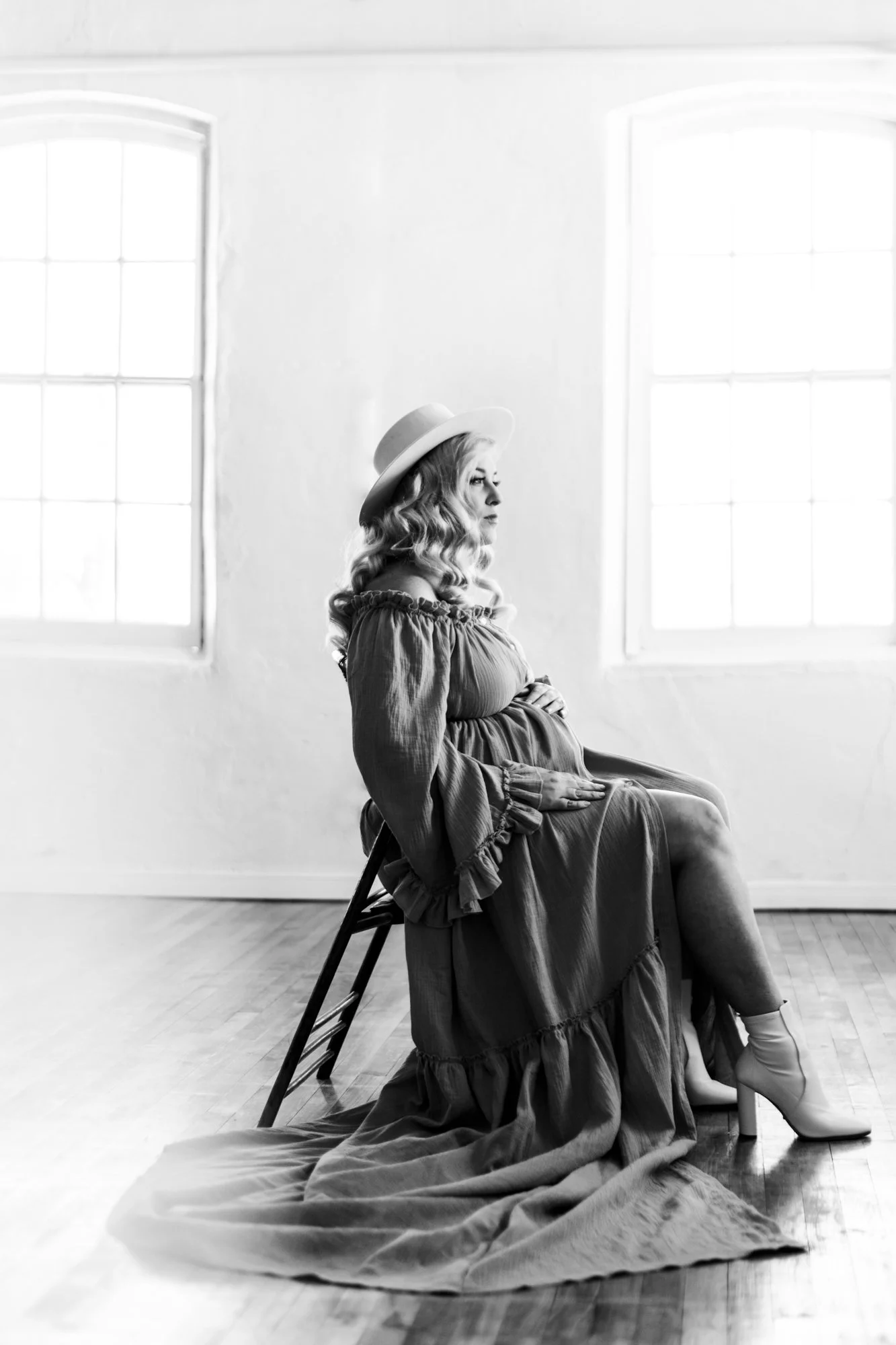 Pregnant woman in a flowing dress sitting on a chair, wearing a wide-brimmed hat, in a bright room with large windows.