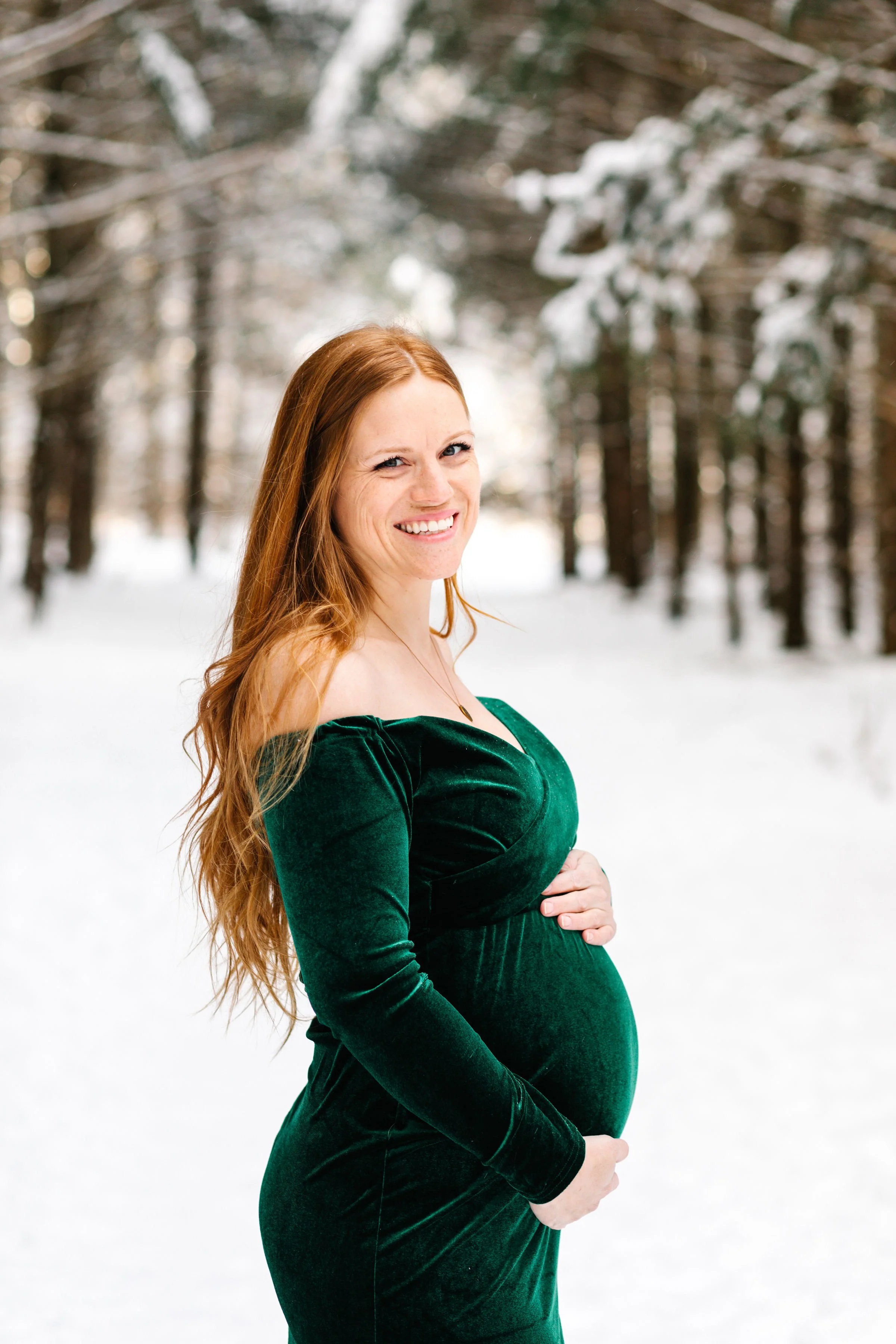 Pregnant woman in a green dress smiling in a snowy forest.