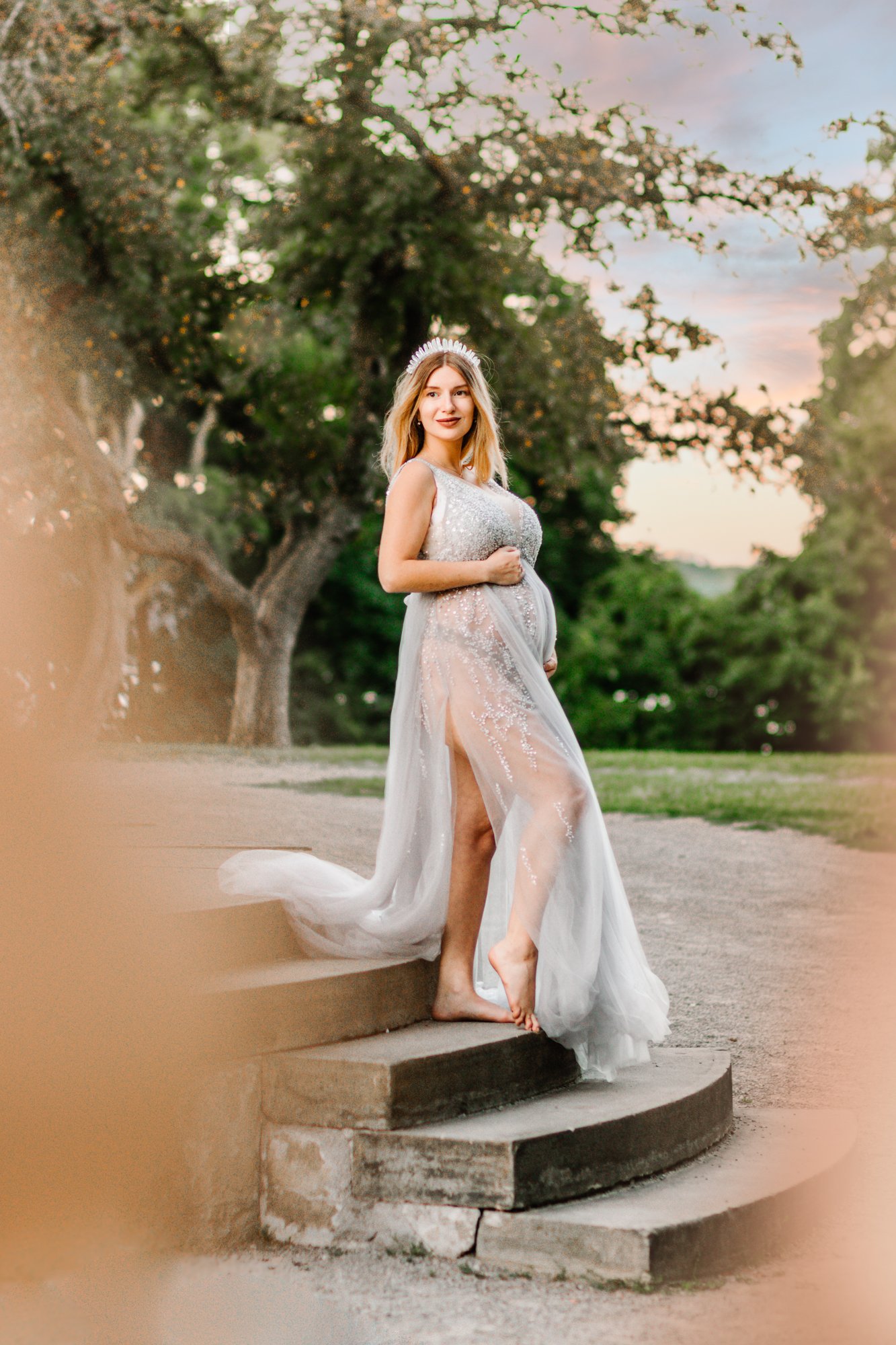 Pregnant woman in a sparkling gown with a crown, standing outdoors on steps with trees in the background.