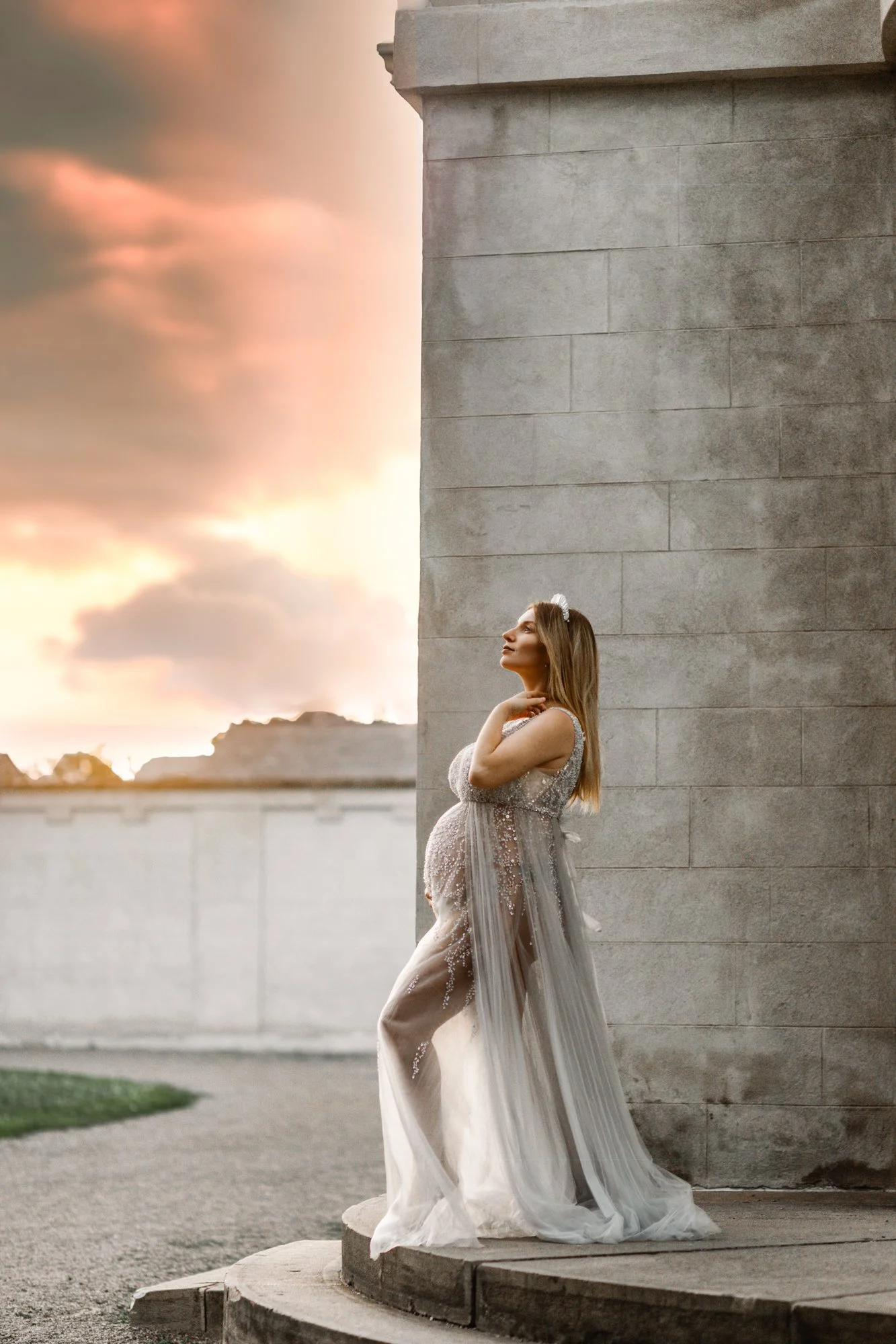 Pregnant woman in a sheer gown posing against a stone structure at sunset.