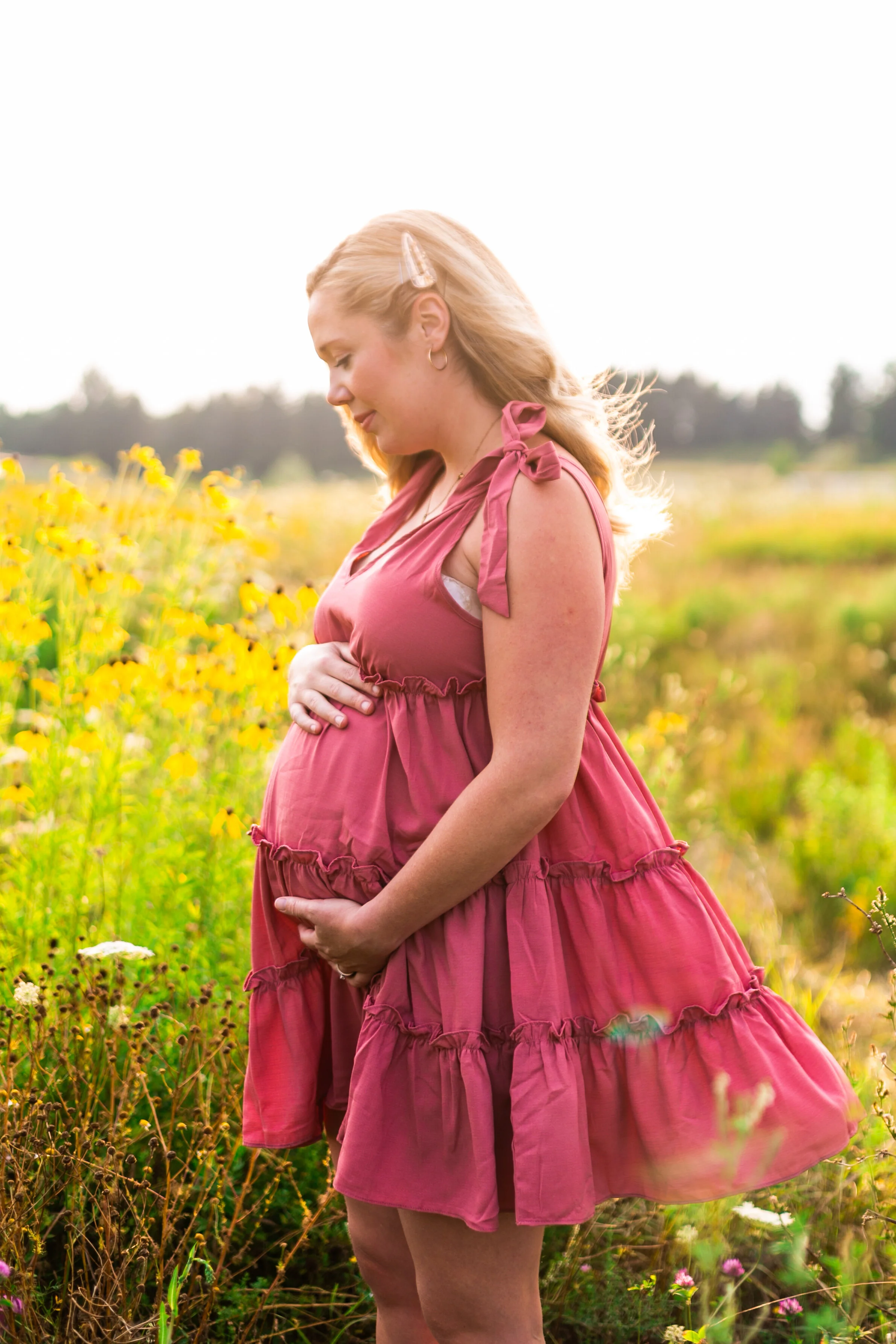 Pregnant woman in pink dress holding her belly in a field of yellow flowers.