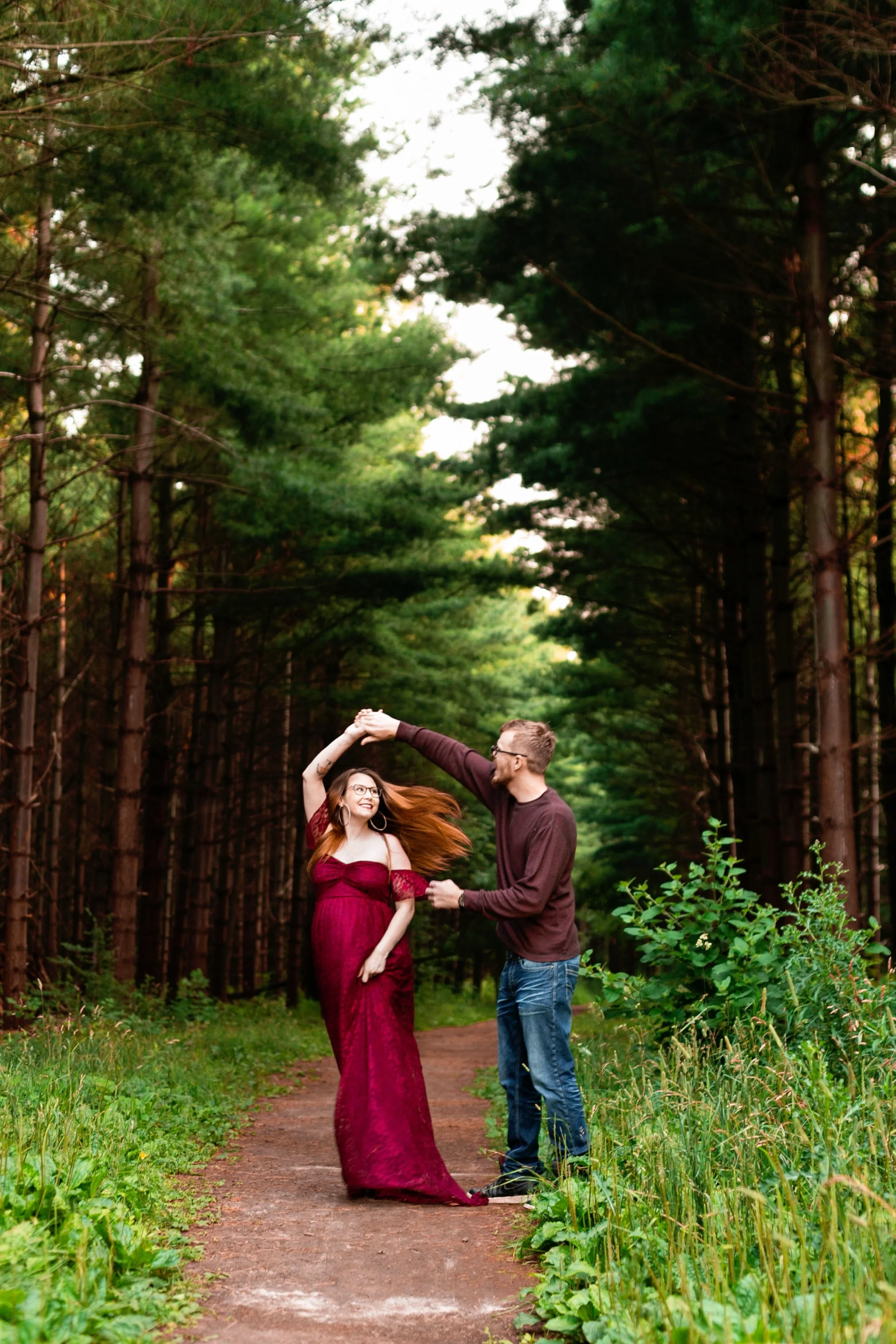 A couple dancing in a forest path, with the woman in a long red dress and the man in casual attire, surrounded by tall green trees.