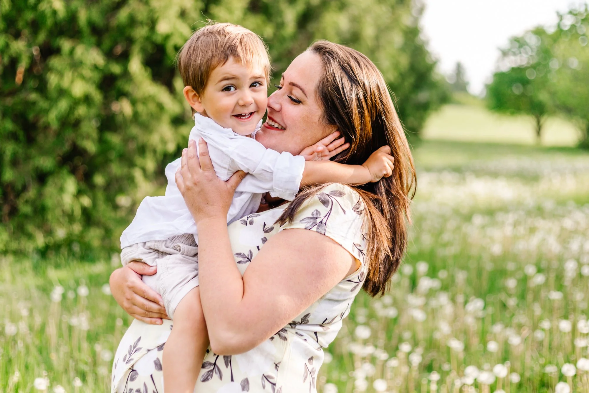 Mother holding child in a green field with dandelions, both smiling.
