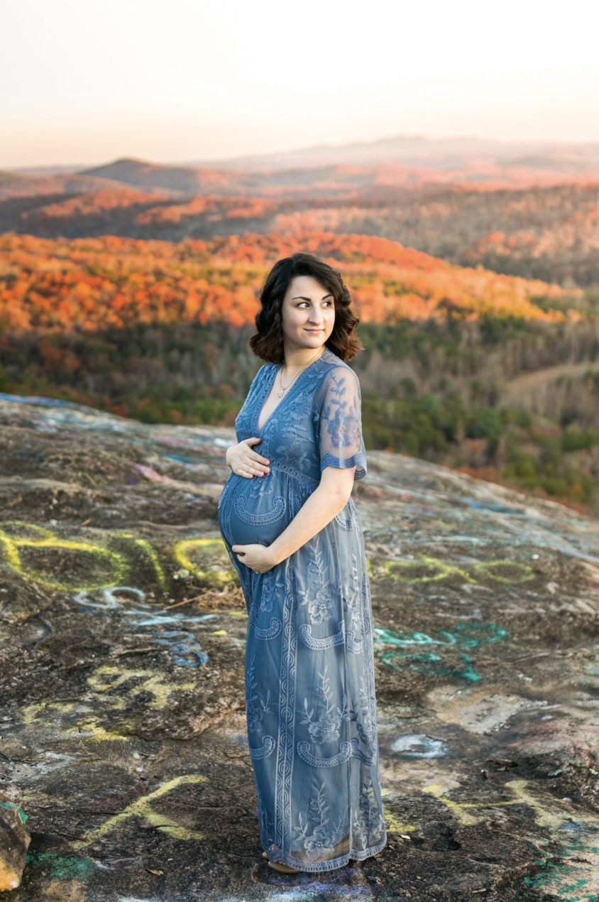 Pregnant woman in a blue dress standing on a rock with colorful autumn foliage in the background.