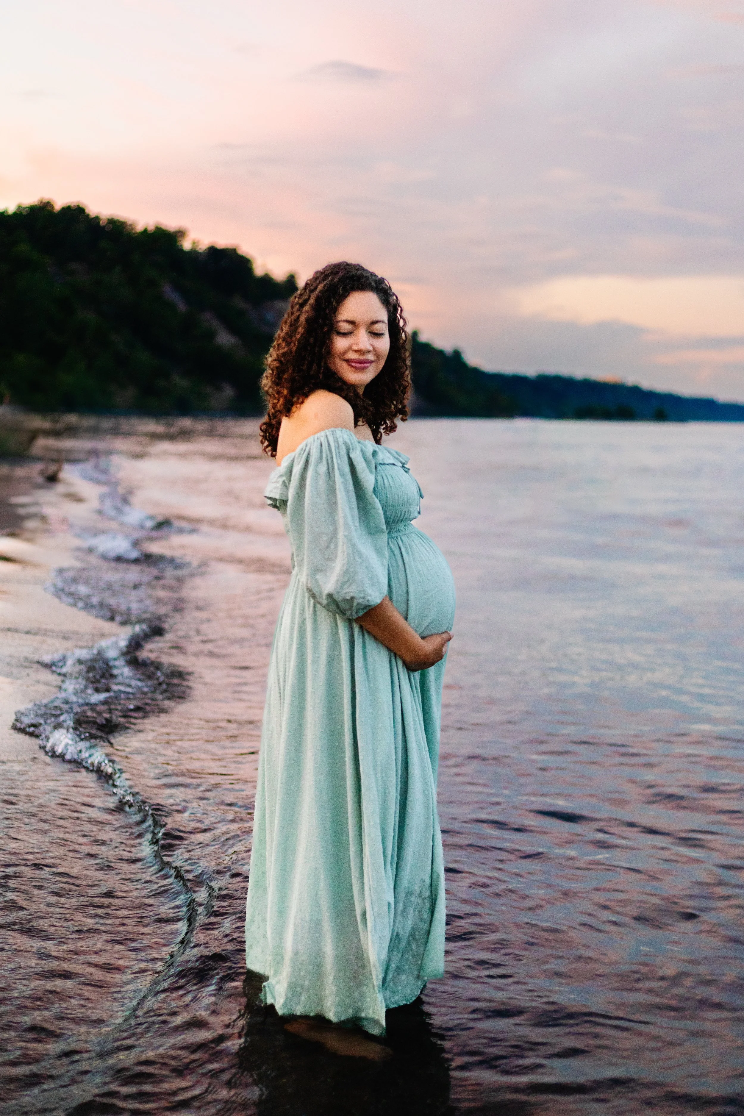 Pregnant woman in a light blue dress standing by the shoreline during sunset.