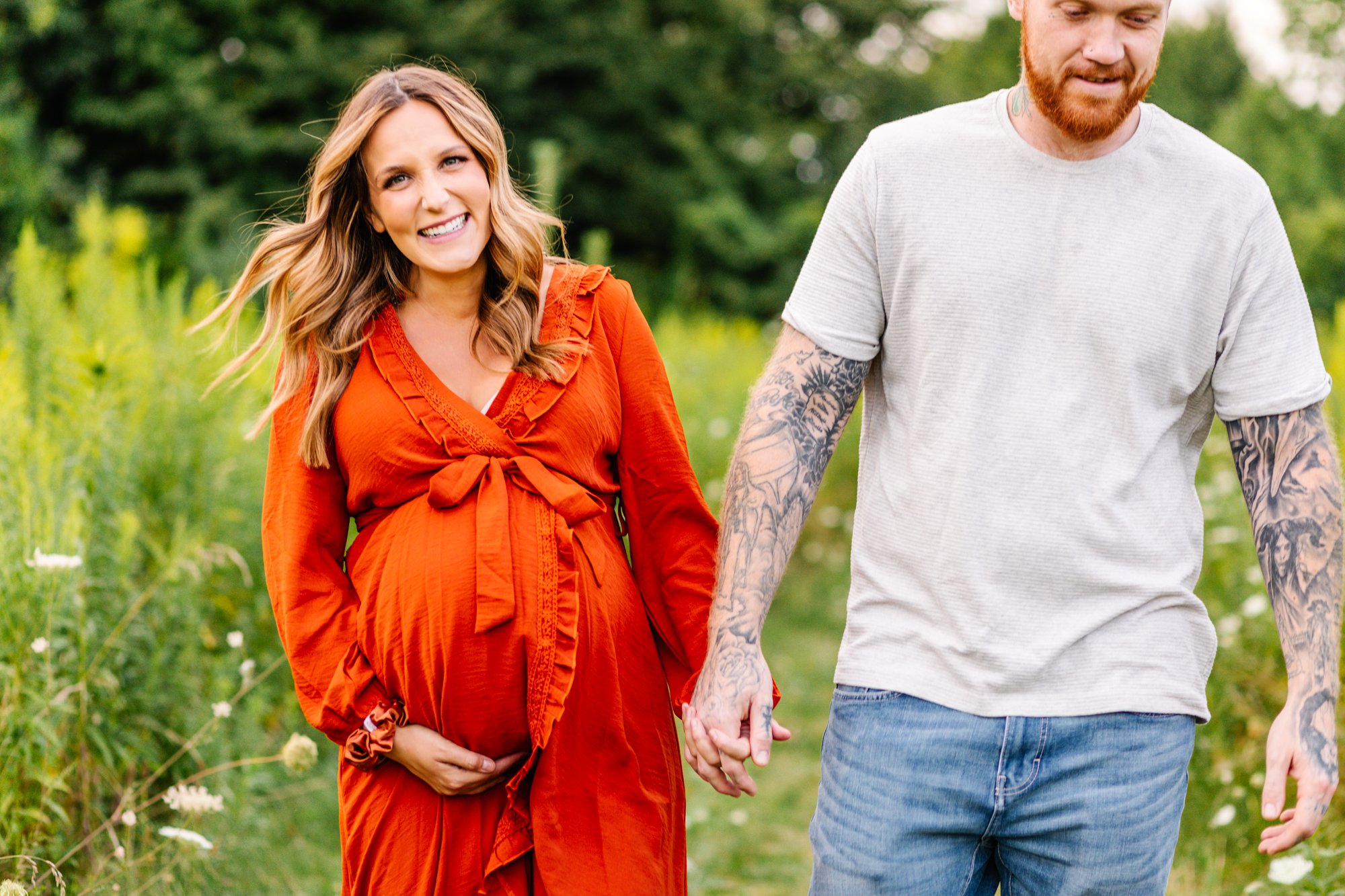 Pregnant woman in an orange dress holding hands with a tattooed man in a field.