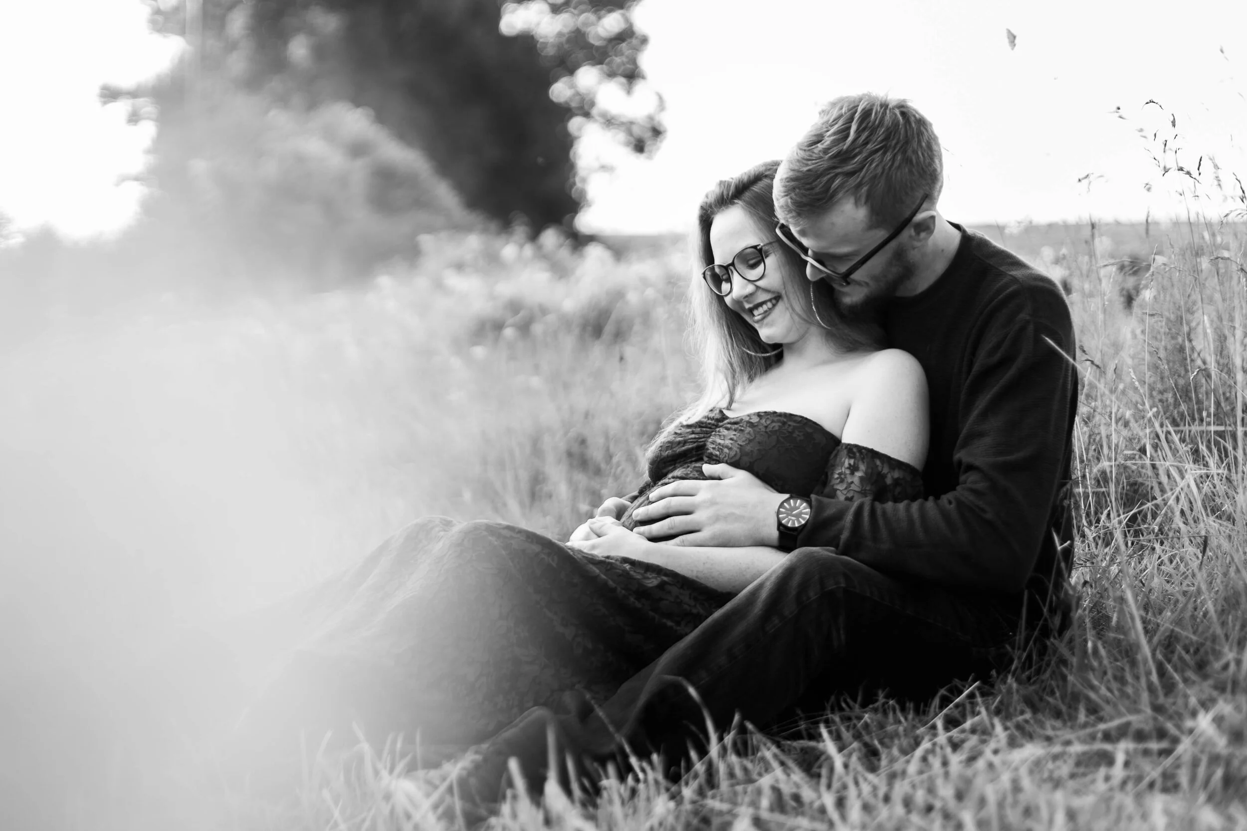 Pregnant couple sitting in a field, smiling at each other, man hugging woman from behind.