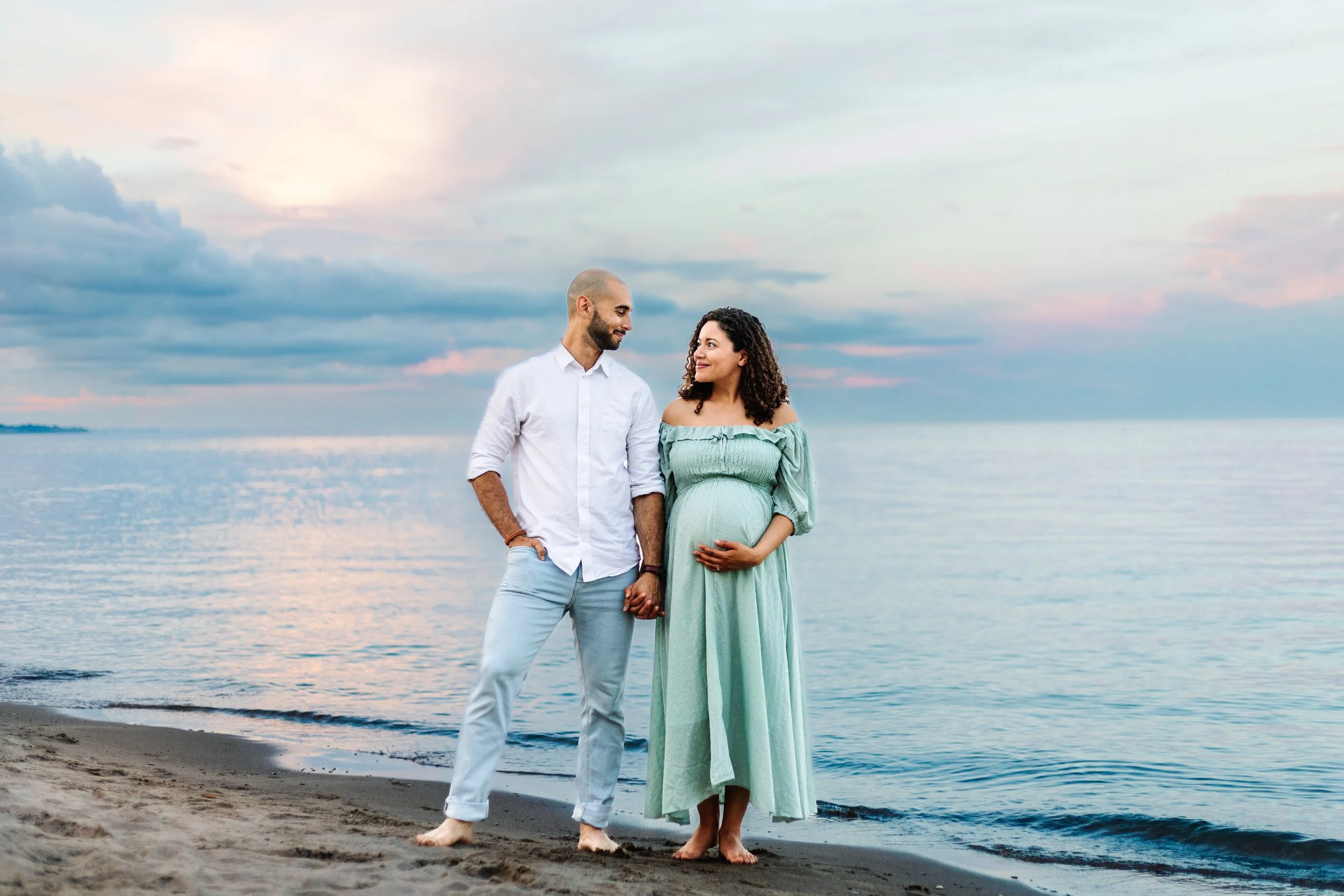 A couple standing on a beach holding hands, with the woman in a green dress and pregnant, during sunset.