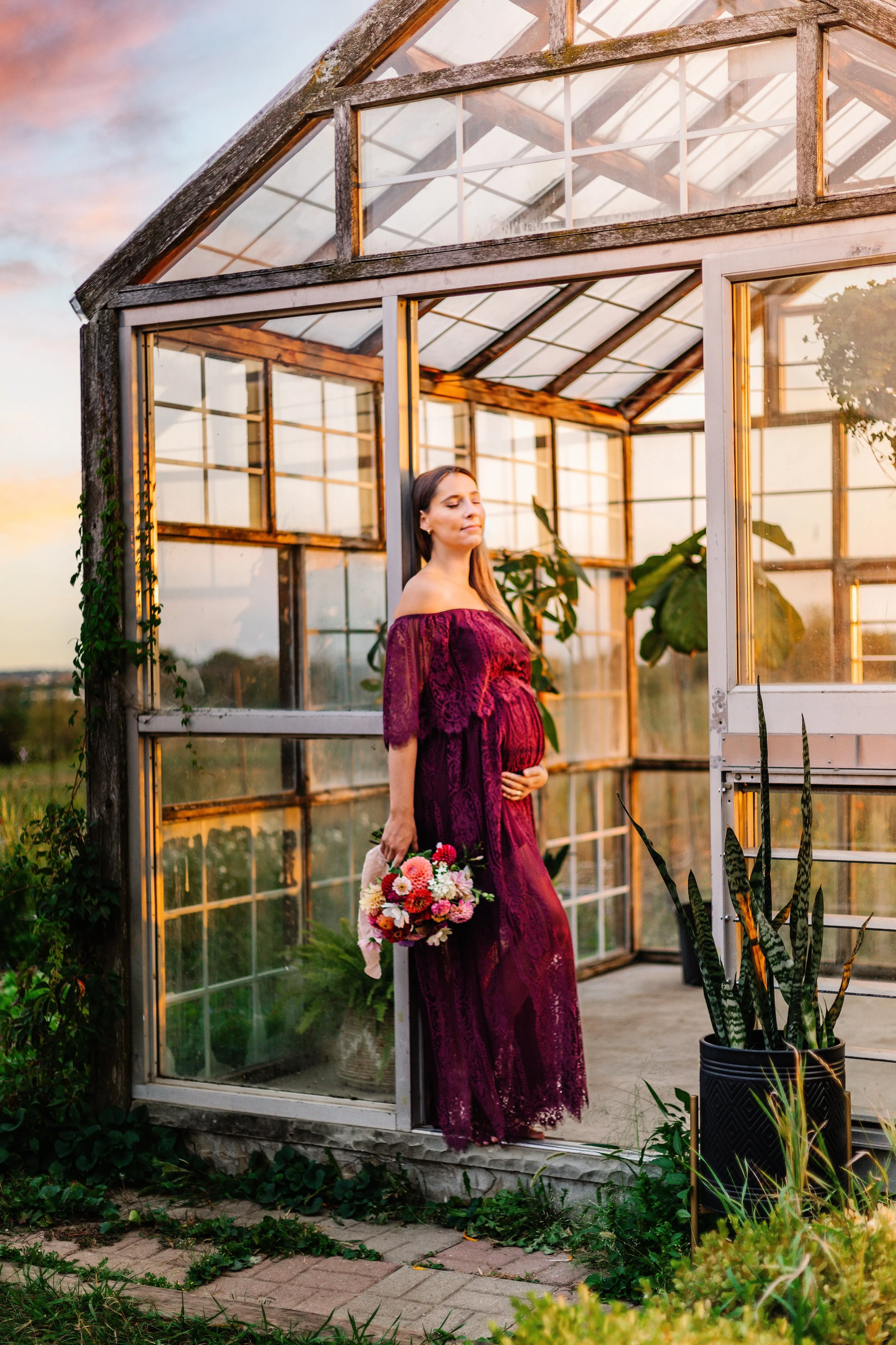 Pregnant woman in a maroon dress holding flowers, stands in front of a greenhouse.