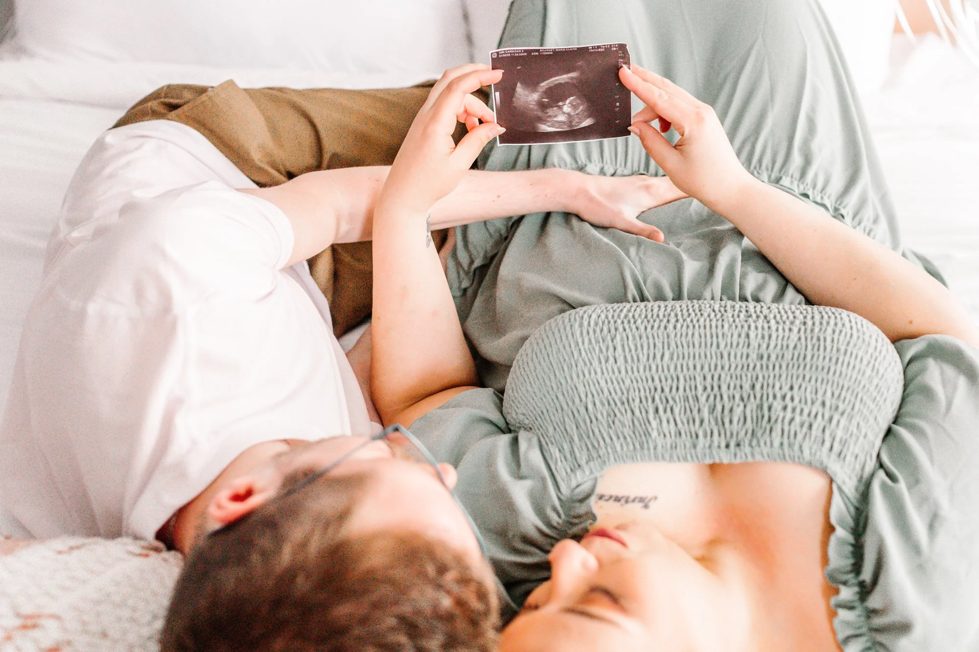 A couple lying on a bed, looking at an ultrasound image. The woman is wearing a green dress, and they both appear content as they hold and gaze at the sonogram together.