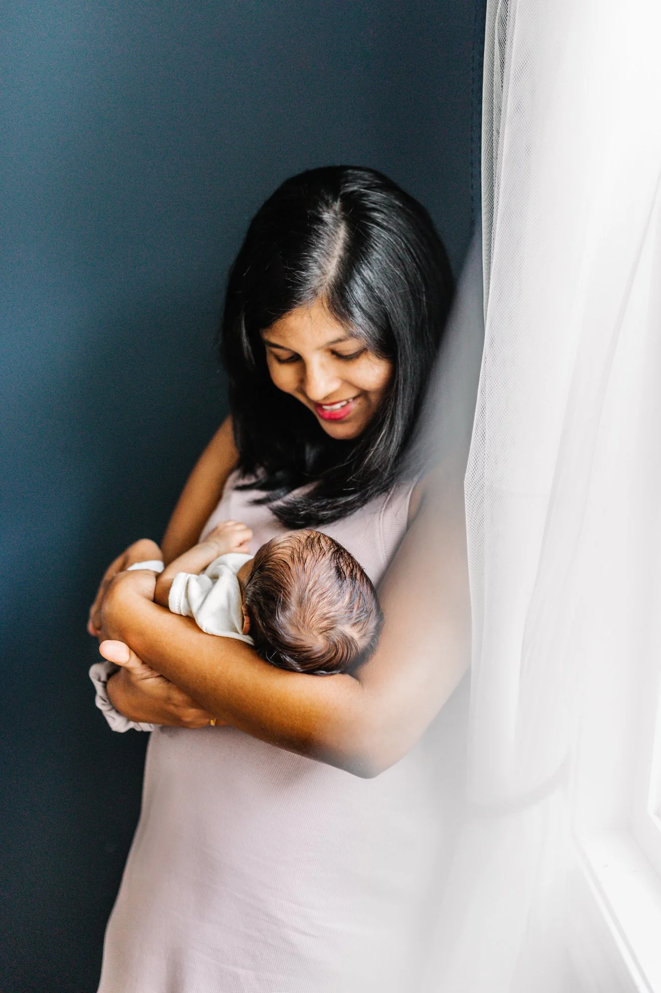 A woman smiling while holding a baby wrapped in a blanket, standing near a window with sheer curtains in soft lighting.