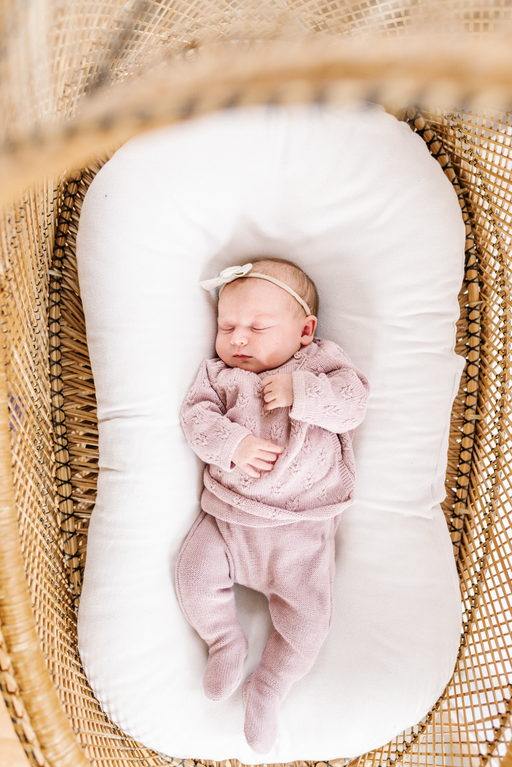 Newborn baby sleeping in a wicker bassinet, wearing a pink outfit and headband.
