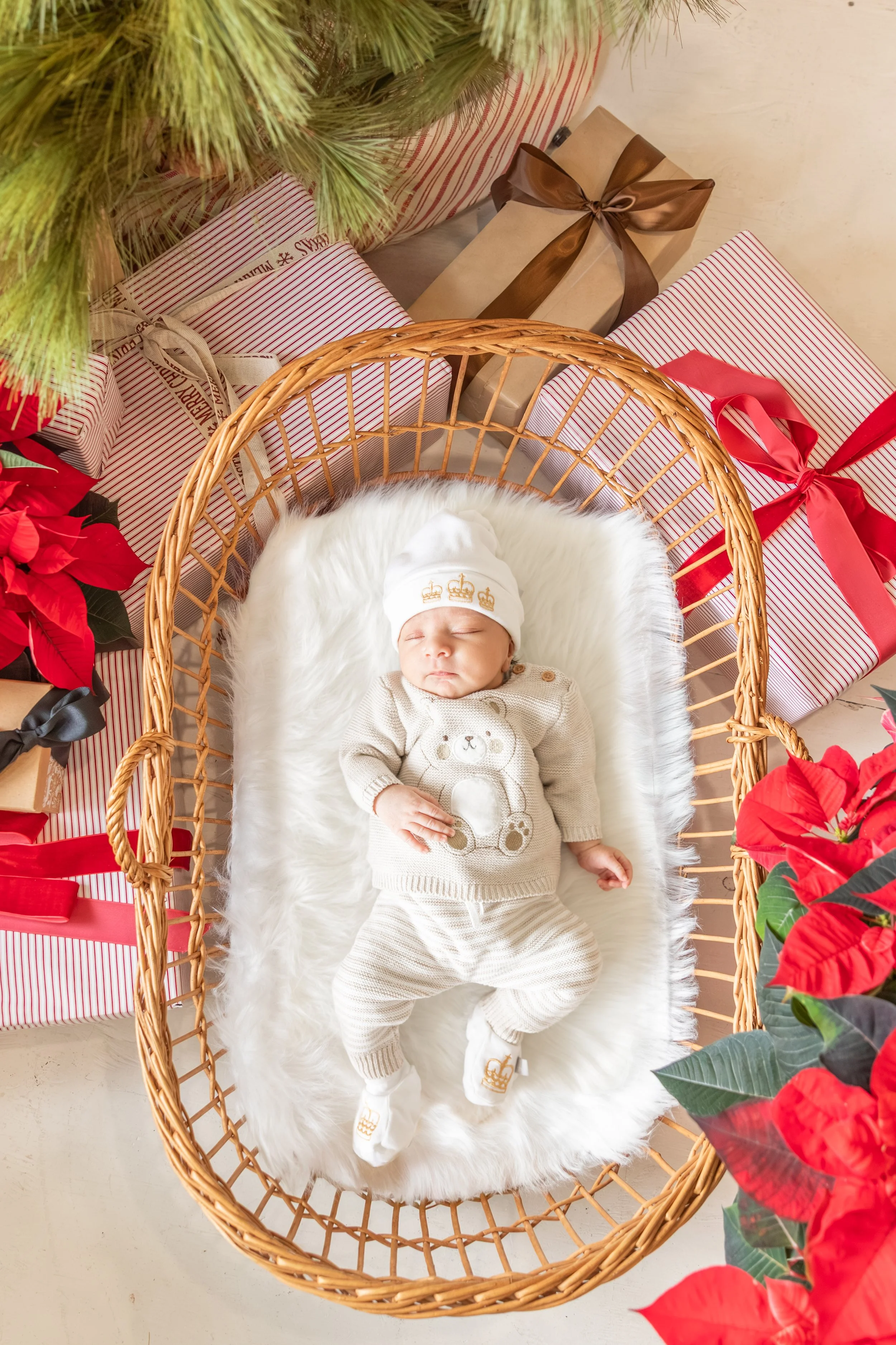 A baby sleeping in a wicker bassinet surrounded by wrapped Christmas presents, poinsettia flowers, and holiday decorations.