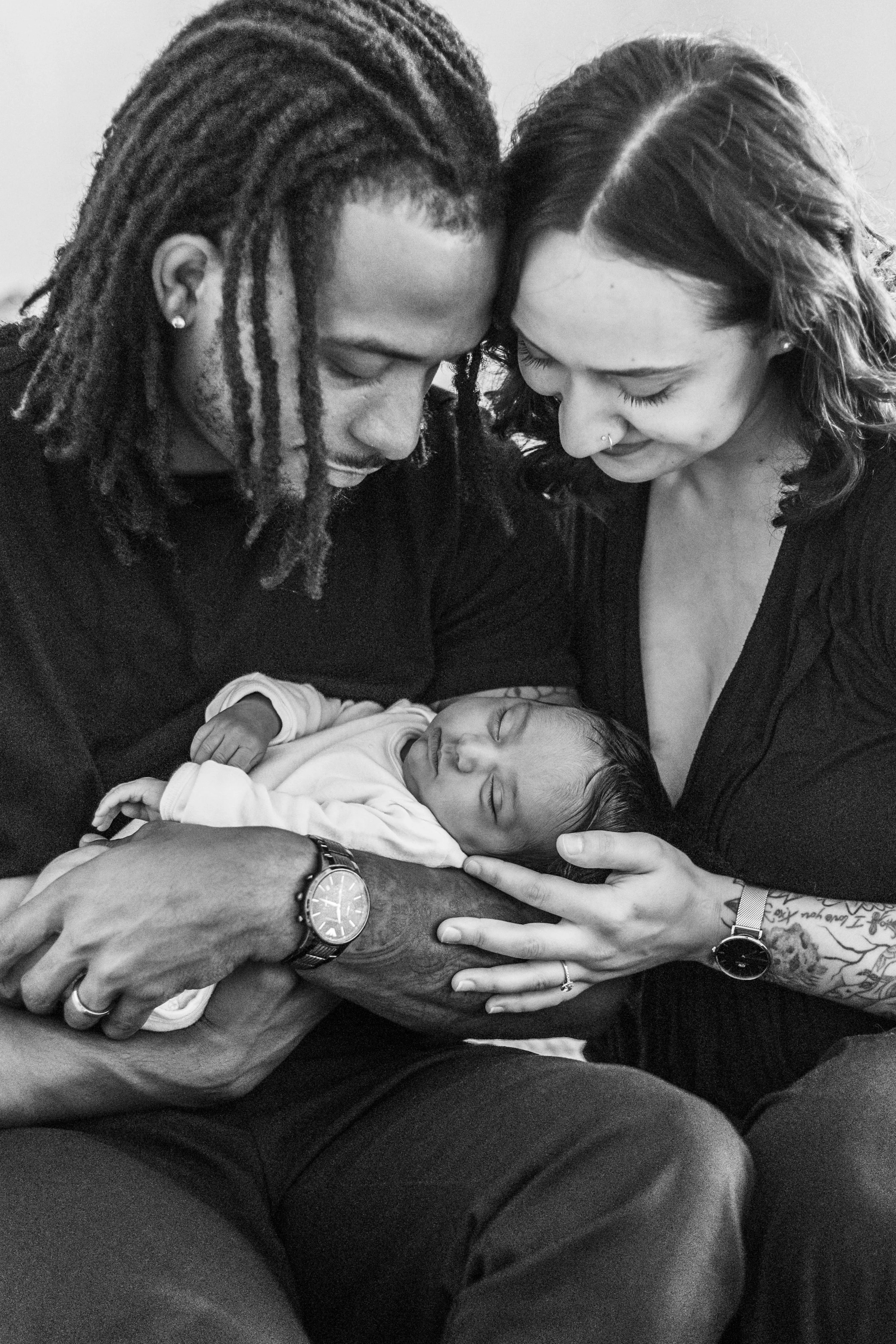 A black and white photo of two adults lovingly holding a sleeping baby between them, both looking down and smiling at the child.