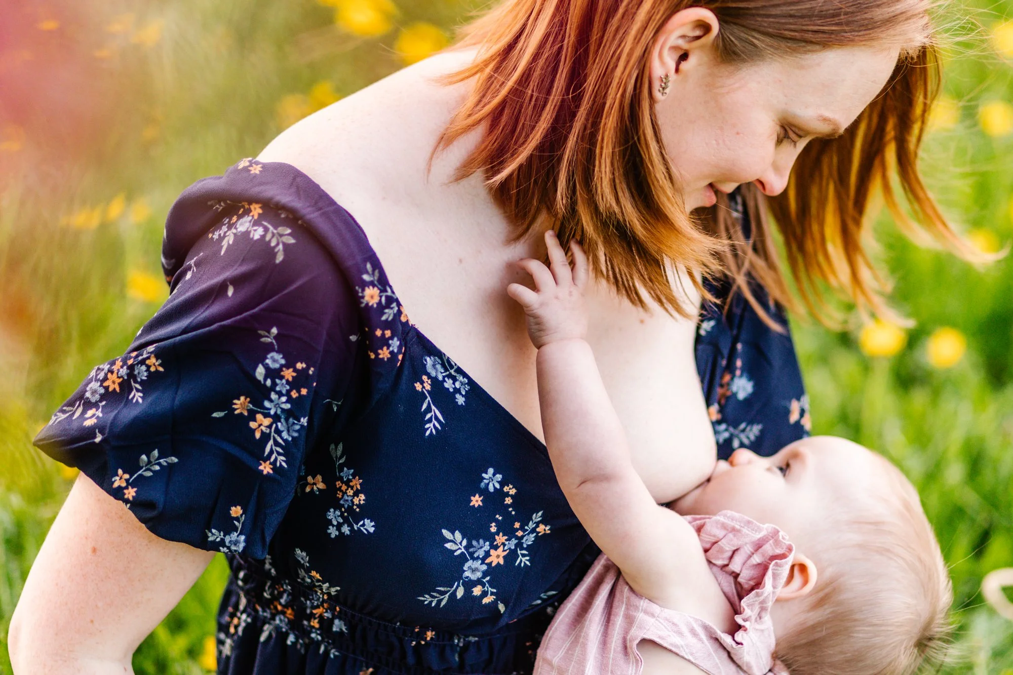 Mother breastfeeding baby outdoors in a field with yellow flowers.