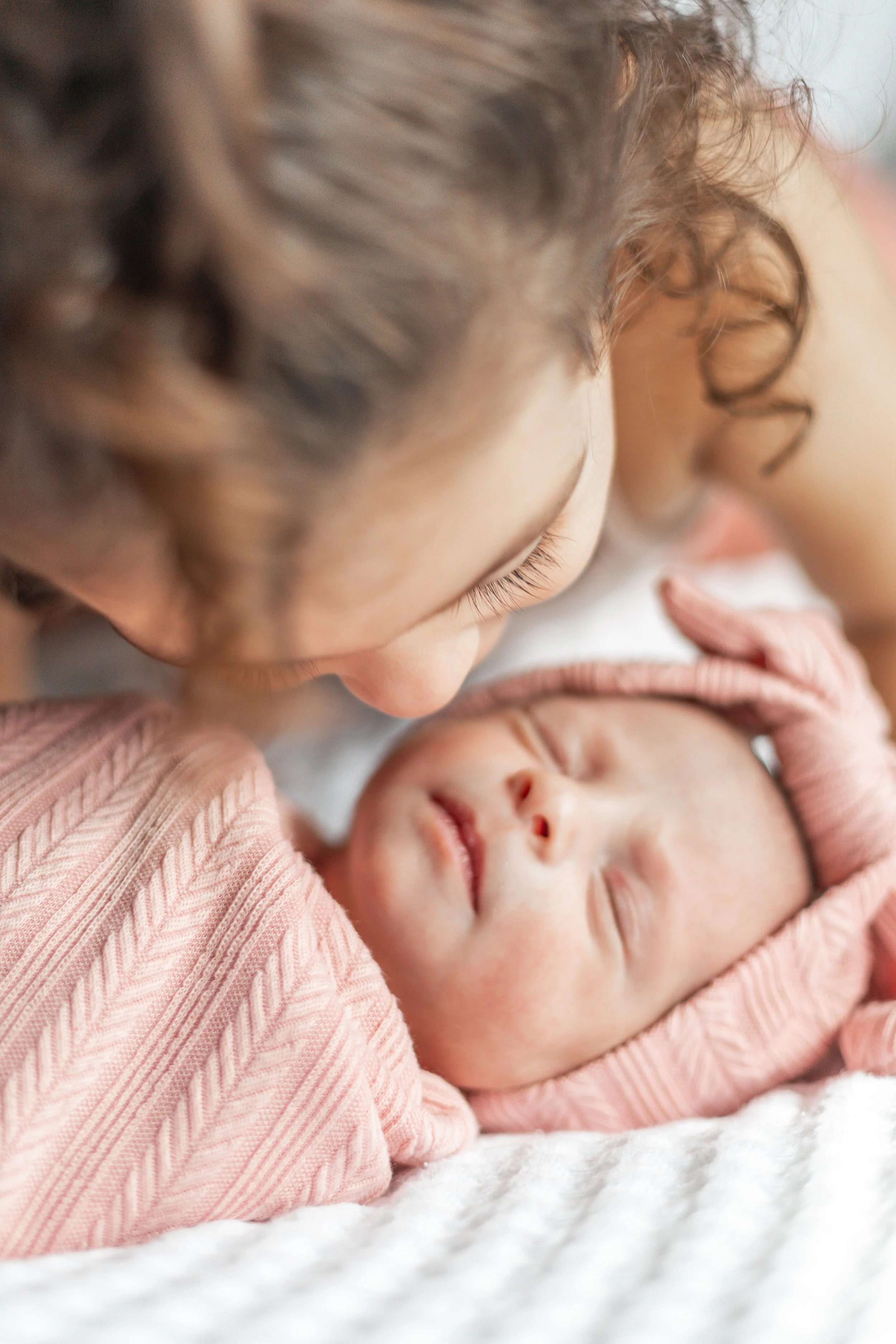 A young child gently kissing a sleeping baby wrapped in a pink blanket and hat, lying on a soft white surface.