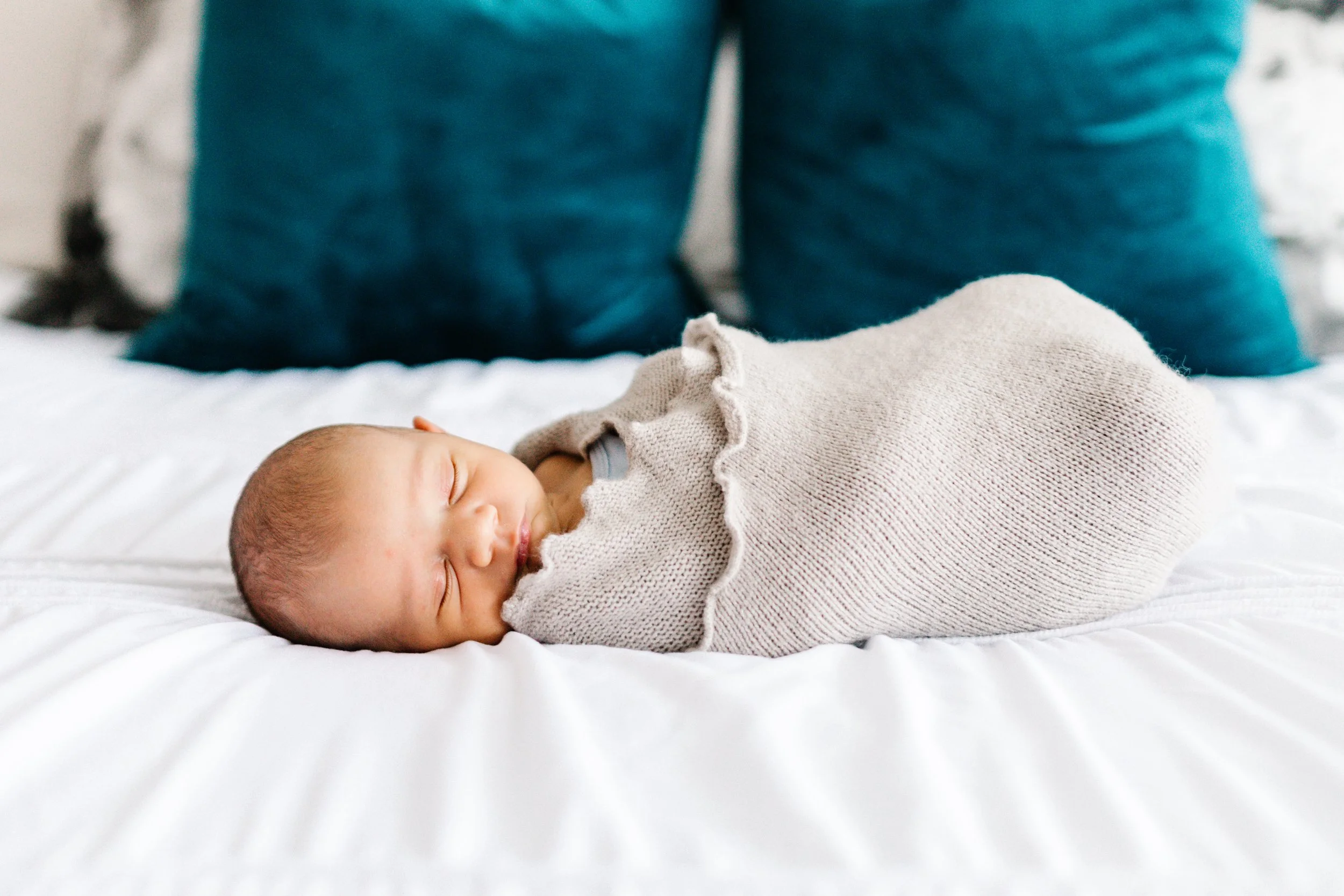 Newborn baby sleeping on white bed, wrapped in a beige knit blanket with teal pillows in the background.