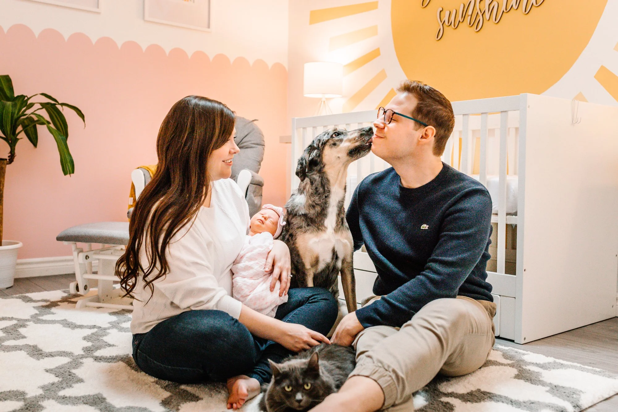 A family sitting in a nursery with a pink and yellow theme. The mother holds a newborn baby, the father interacts with a grey dog, and a cat sits in front of them on the rug. A white crib is in the background.