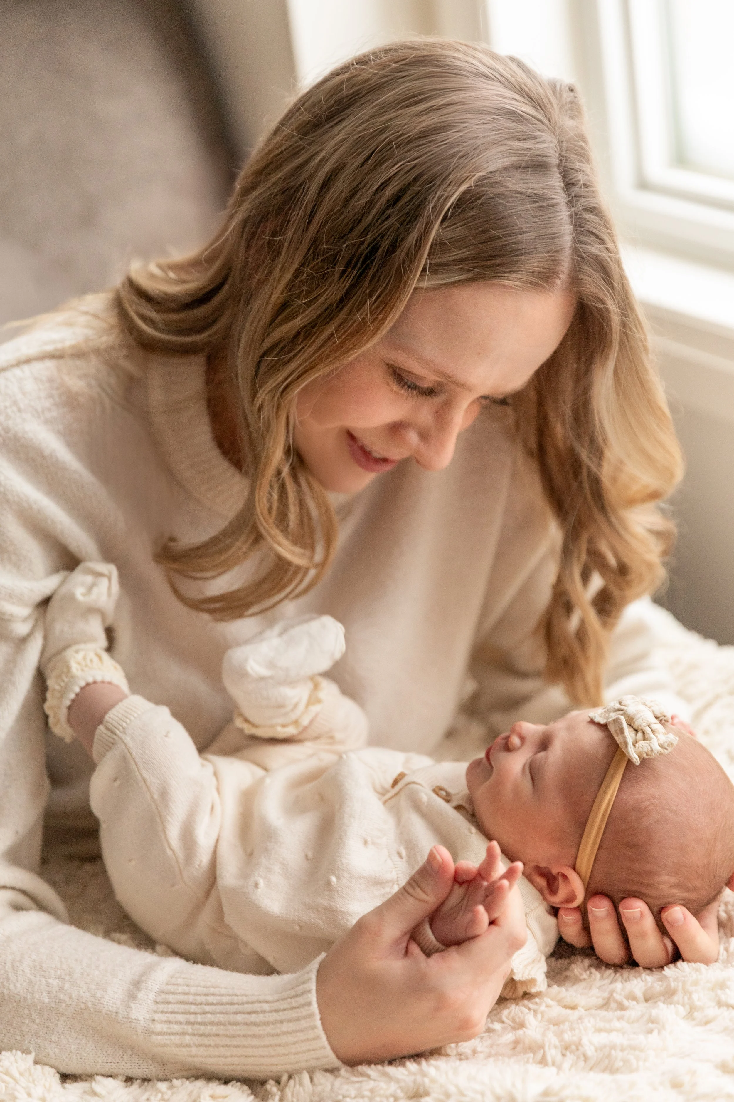 A woman smiling and looking down at a baby lying on a soft blanket, the woman holding the baby's hand.