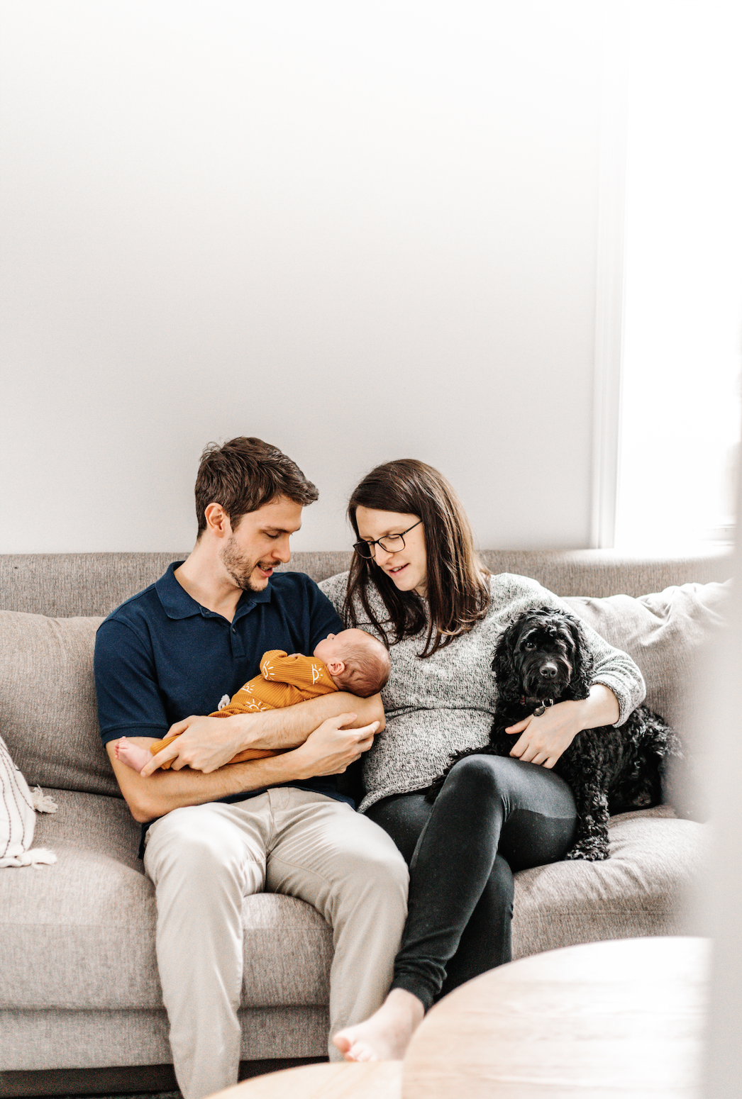 A couple sitting on a beige couch with a baby and a black dog; the man is holding the baby.