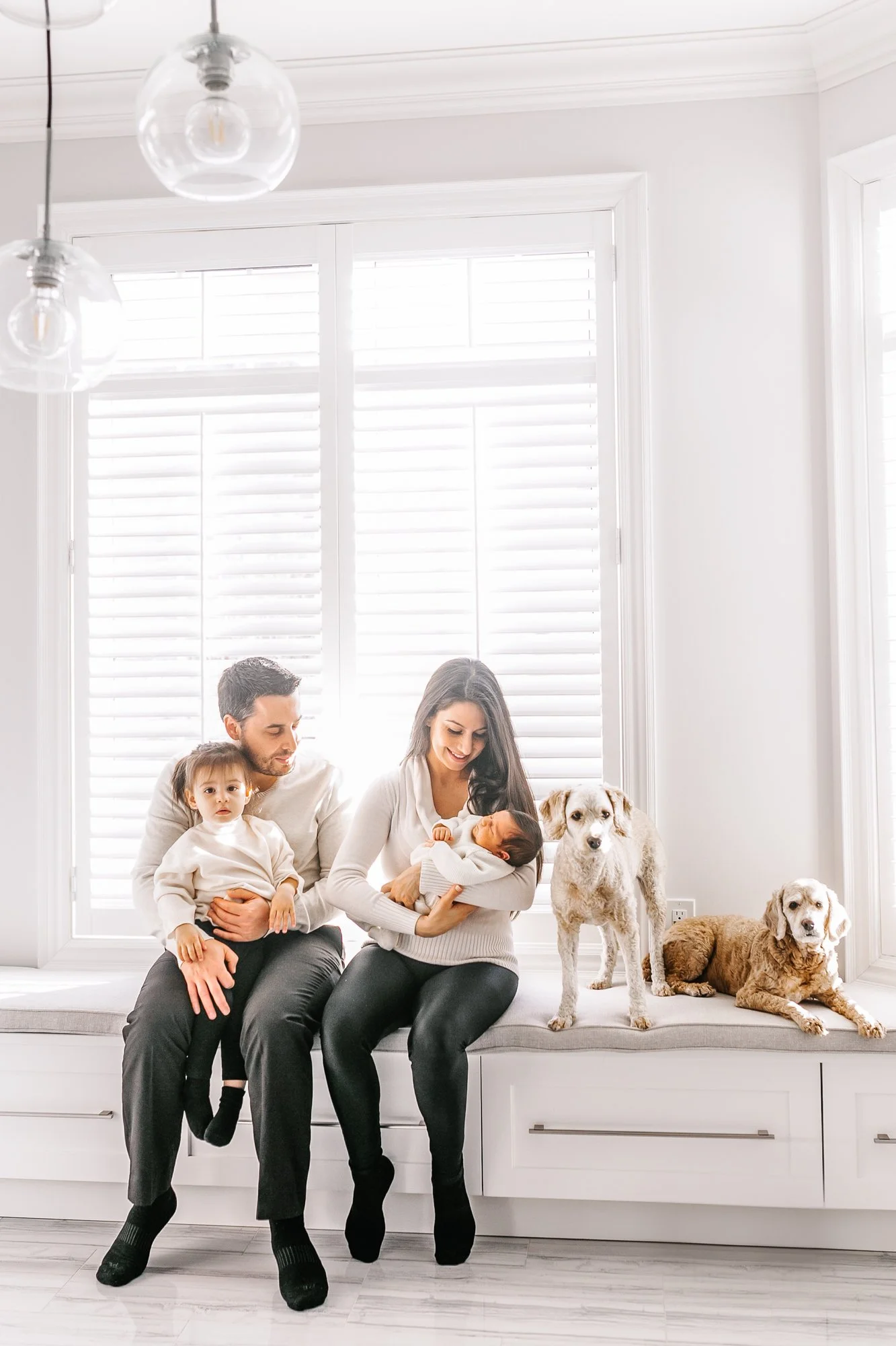 A family sitting together on a bench in a bright room with white shutters. The father is holding a child, while the mother holds a baby. Two dogs are sitting beside them.