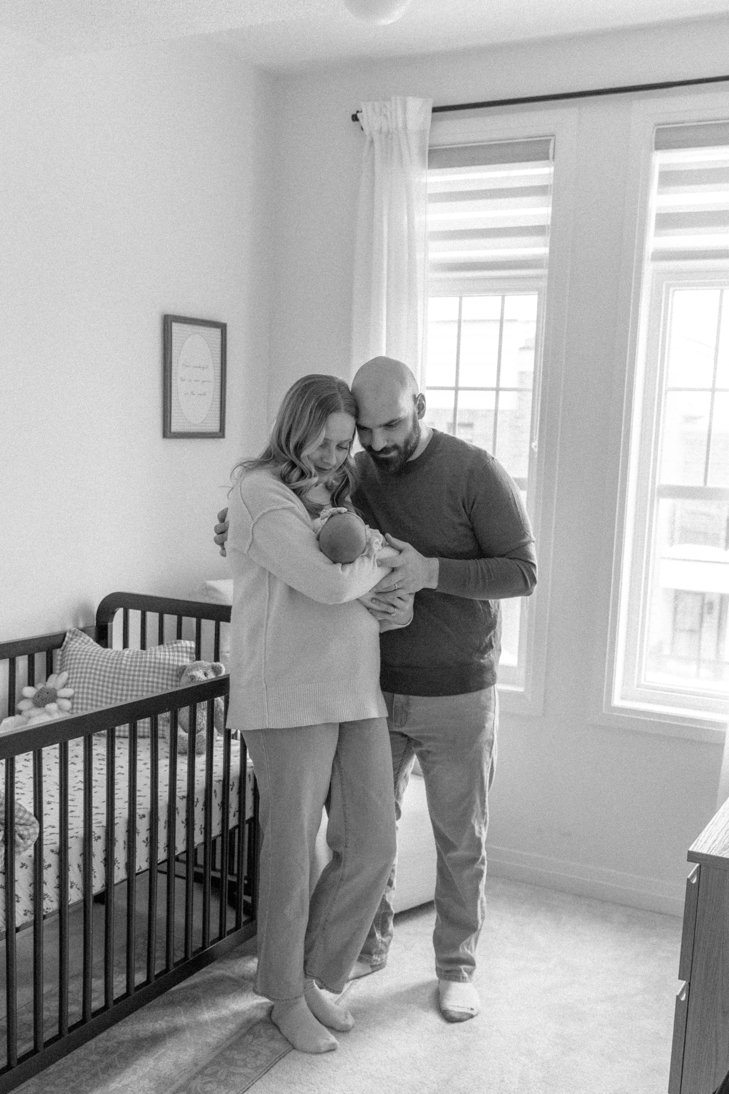 A couple holding a newborn baby inside a bedroom with a crib and large windows, captured in black and white.