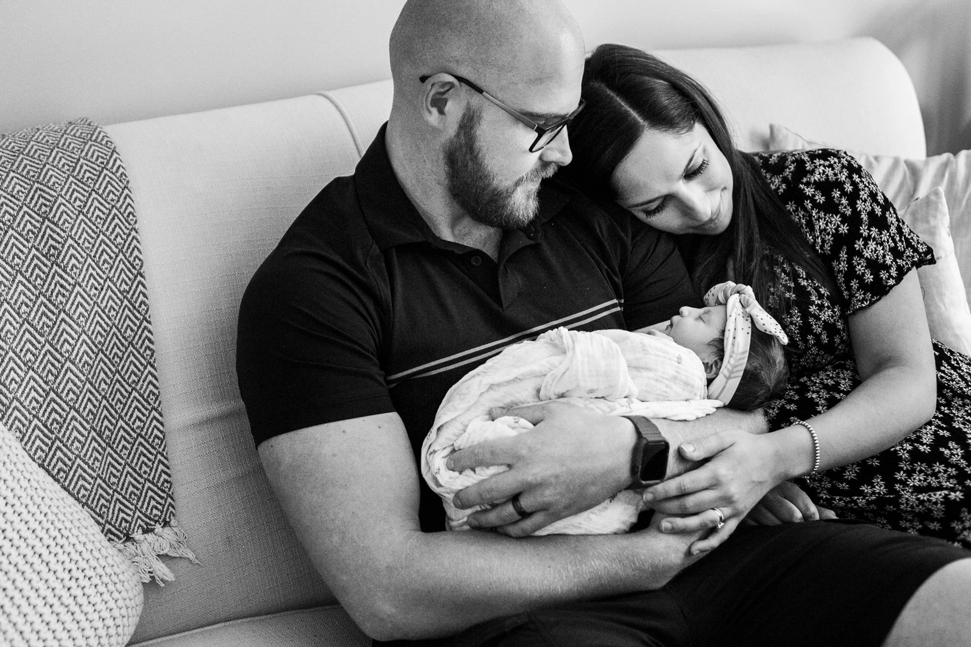 A couple sitting on a couch holding a sleeping baby wrapped in a blanket. The man is wearing a black shirt and glasses, and the woman is wearing a patterned dress. The baby has a headband and is swaddled.