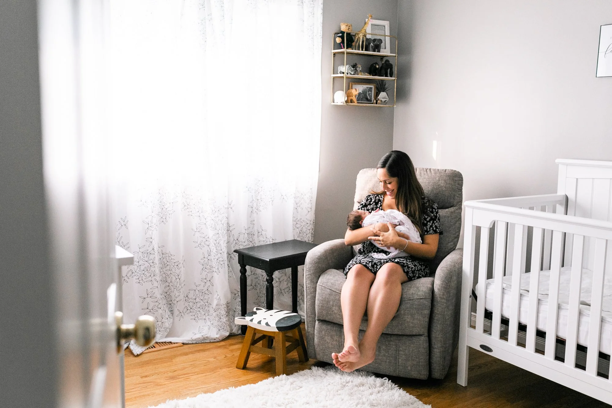 Woman sitting in a gray armchair in a nursery room, holding a baby wrapped in a blanket, next to a white crib. Decor includes a small table, a cushioned footstool, a fluffy rug, and a shelf with toys.