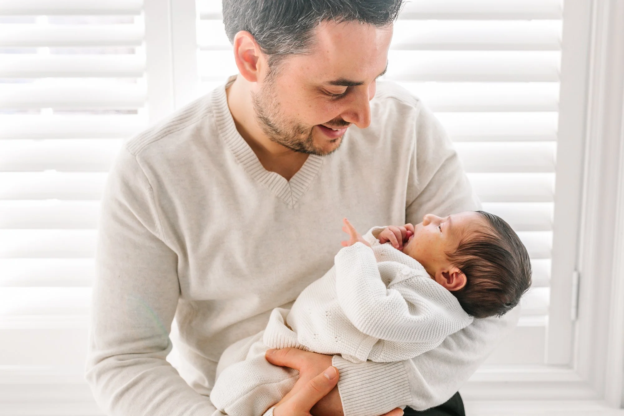 A man holding a baby in a white room with window blinds in the background. The man is smiling at the baby, who is wrapped in a white blanket.