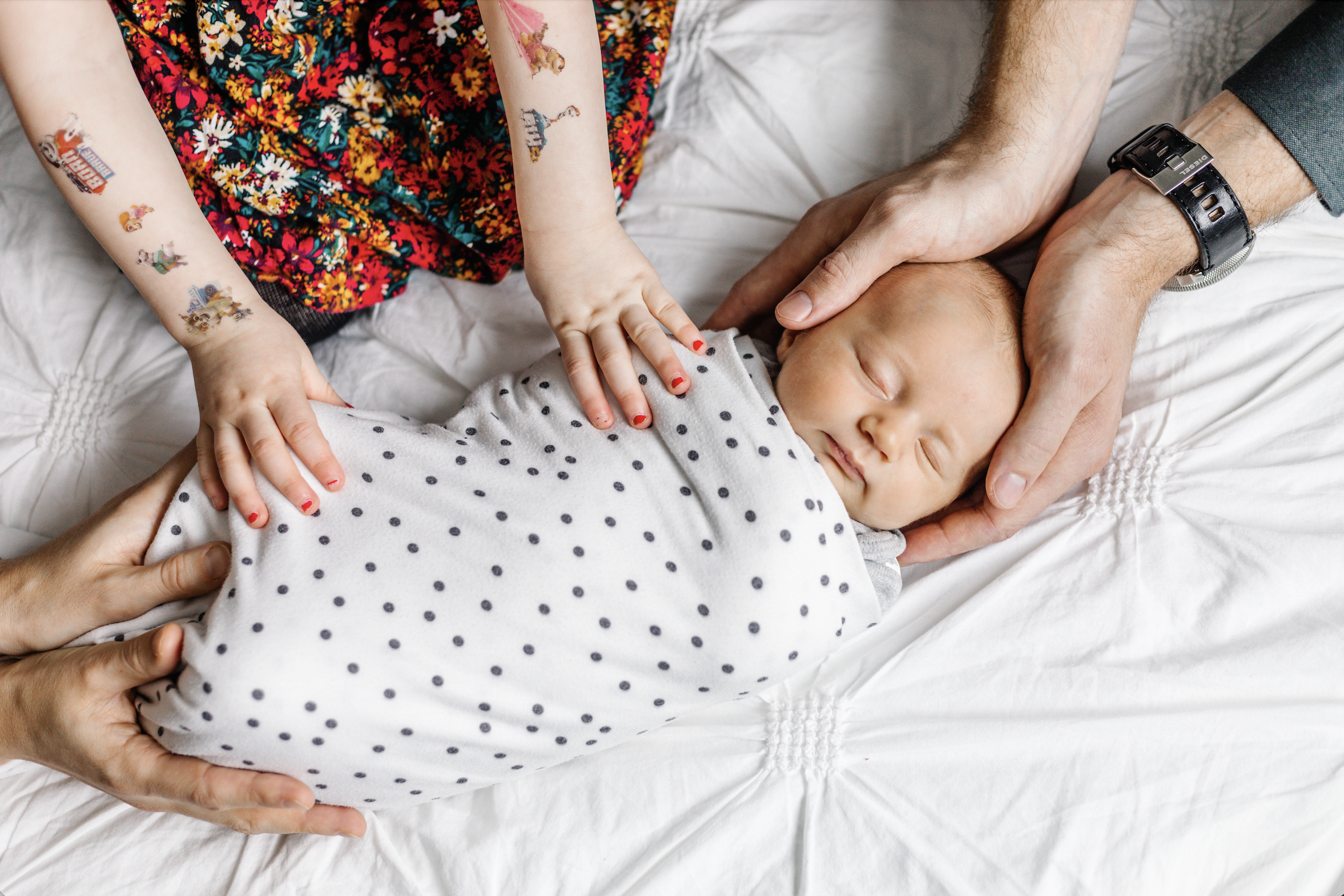 Swaddled newborn sleeping, held by family hands on white blanket, child with temporary tattoos nearby.