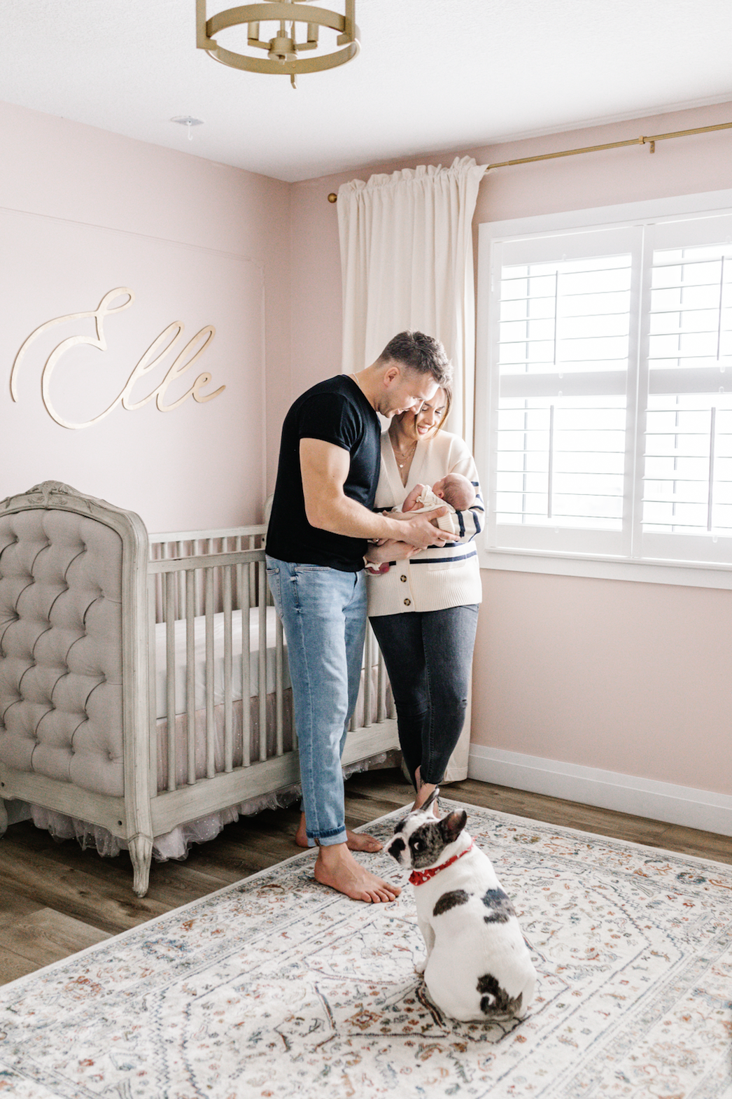 A couple stands in a nursery holding their baby. A crib is in the background with the name 'Elle' on the wall. A dog sits on a patterned rug in the foreground.