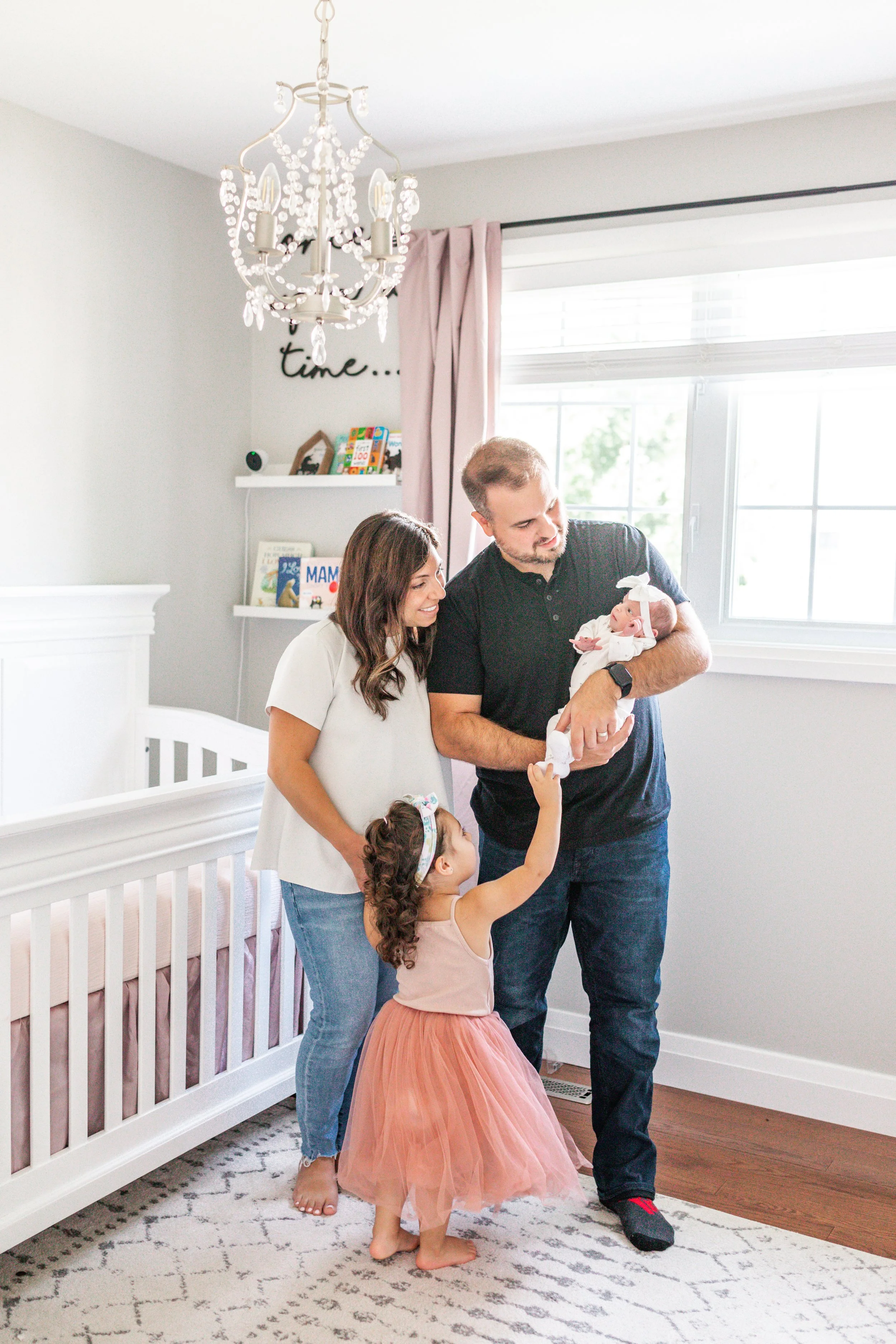 Family of four in a nursery with a white crib, chandelier, and bookshelf.