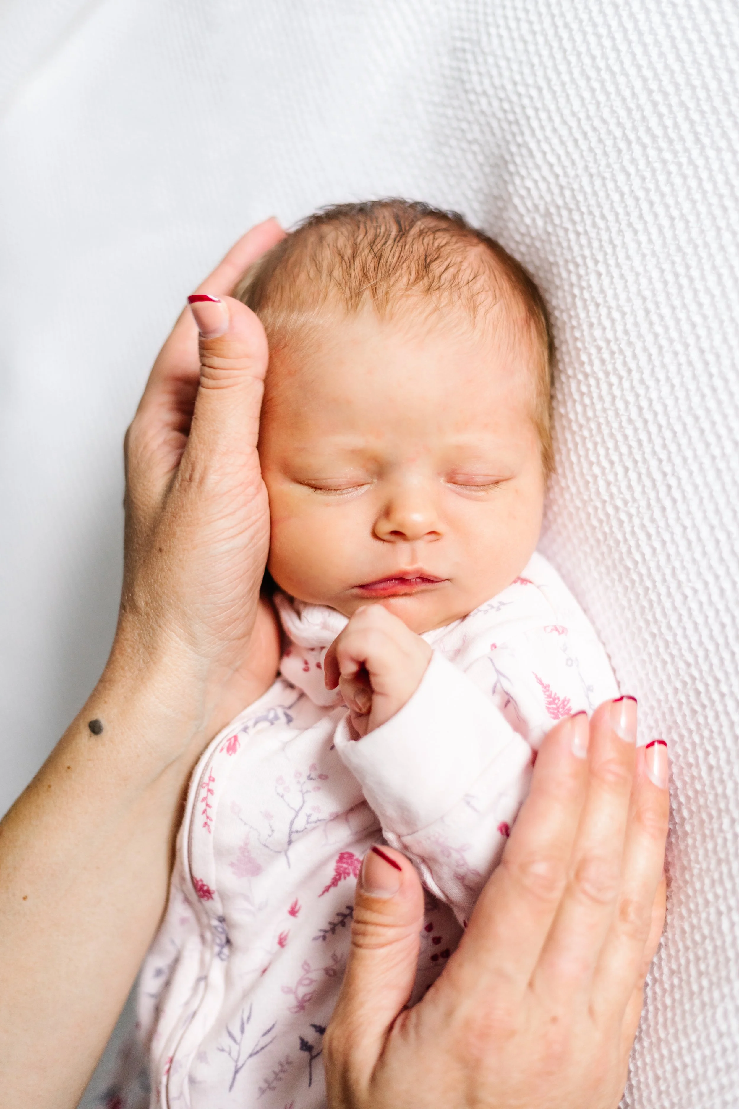 Newborn baby sleeping, held gently by adult hands, wrapped in a floral-patterned onesie.
