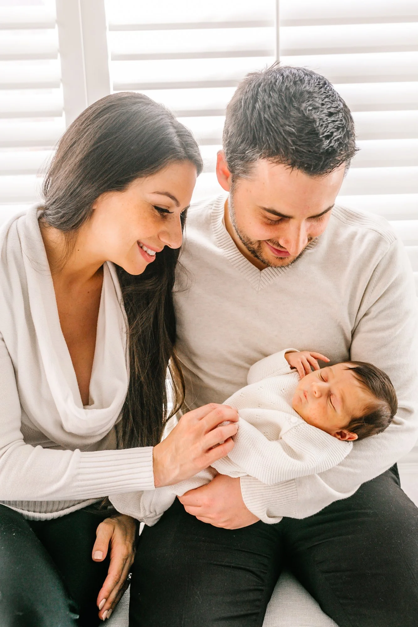 A couple smiling at their newborn baby, who is sleeping in the father's arms. The family is sitting in front of a window with white blinds.