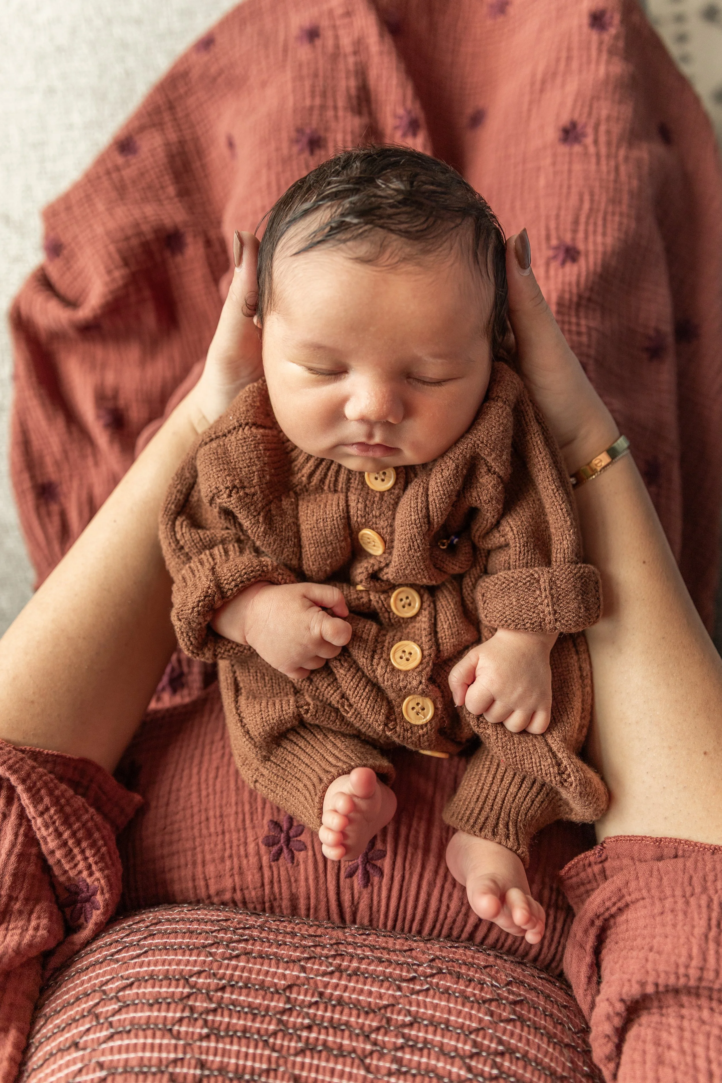 A newborn baby dressed in a brown knitted onesie, sleeping peacefully while being held gently in an adult's hands.