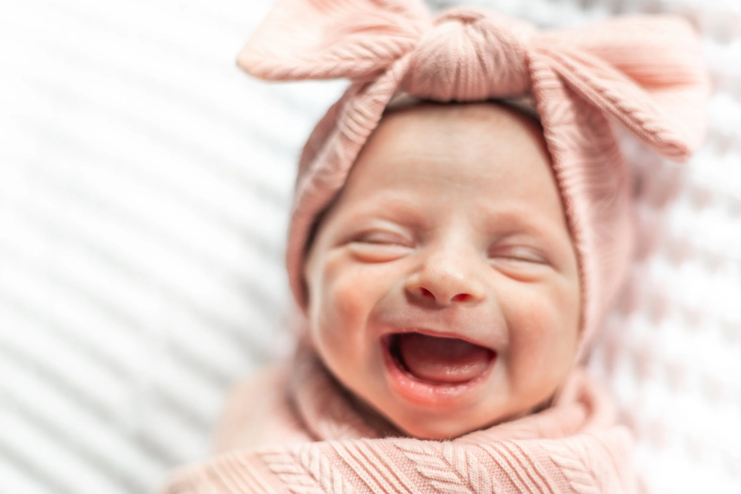 A smiling baby wrapped in a pink blanket wearing a pink headband with a bow.