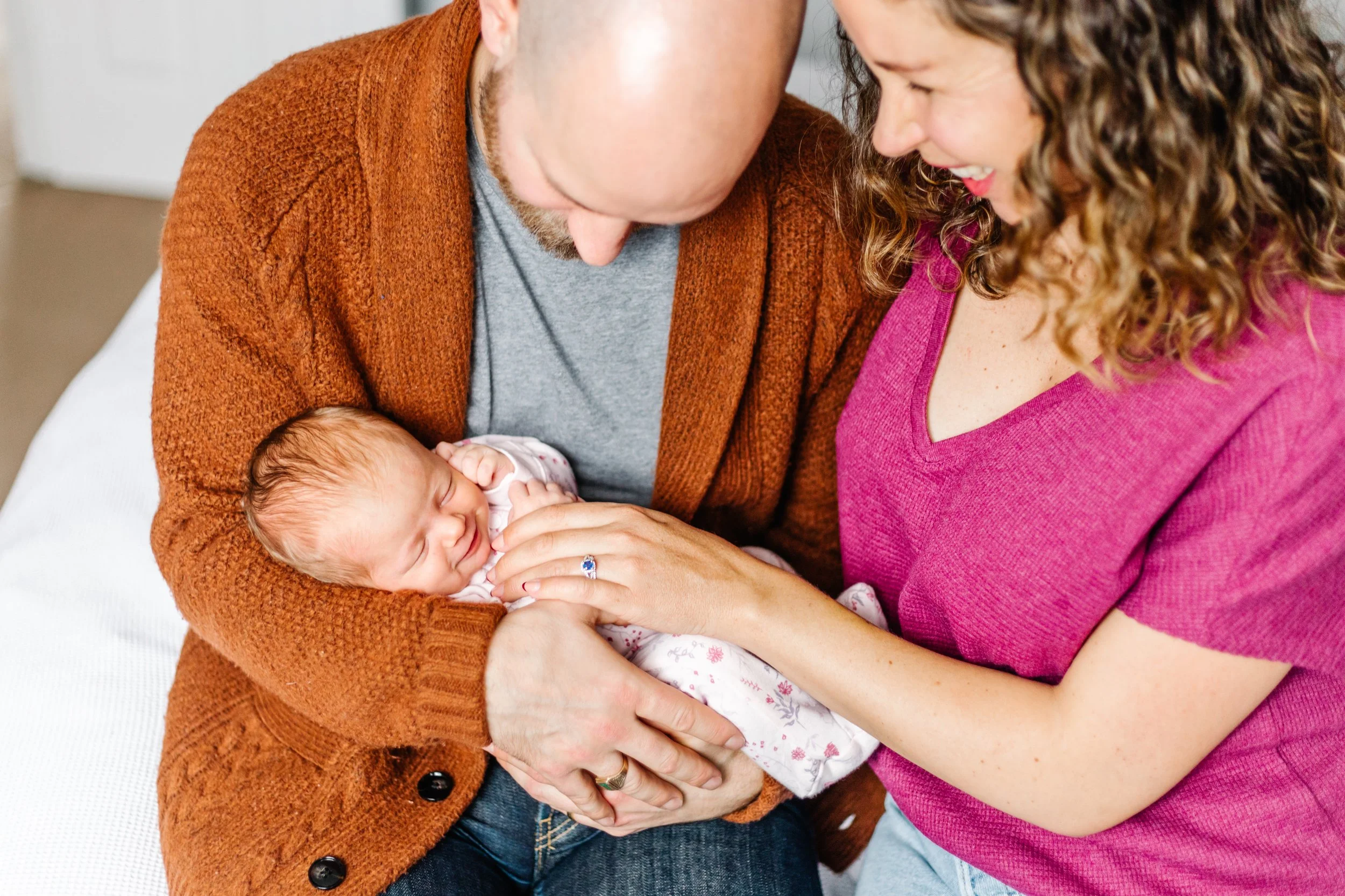 A couple holding a newborn baby wrapped in a patterned blanket. The man is wearing a brown cardigan, and the woman is wearing a pink top. They are looking lovingly at the baby.