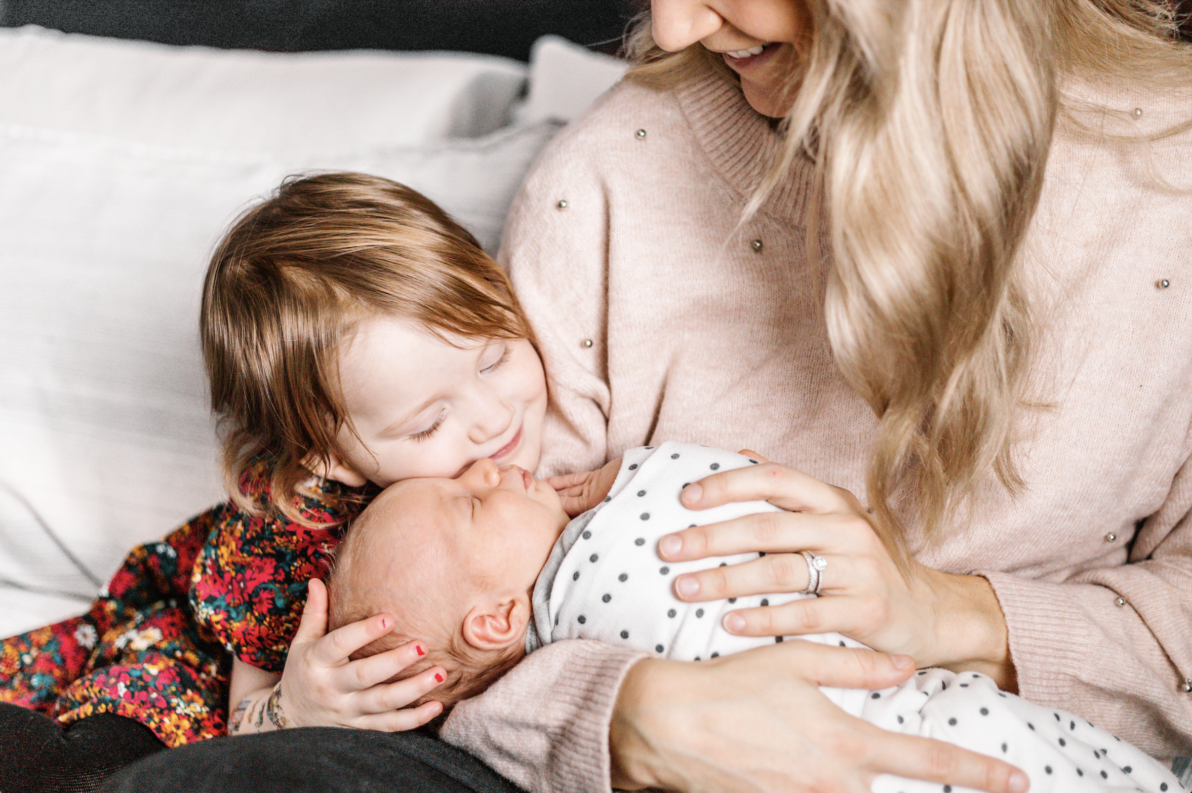 A young child cuddling a newborn baby wrapped in a white blanket with black polka dots, held by a woman in a pink sweater.