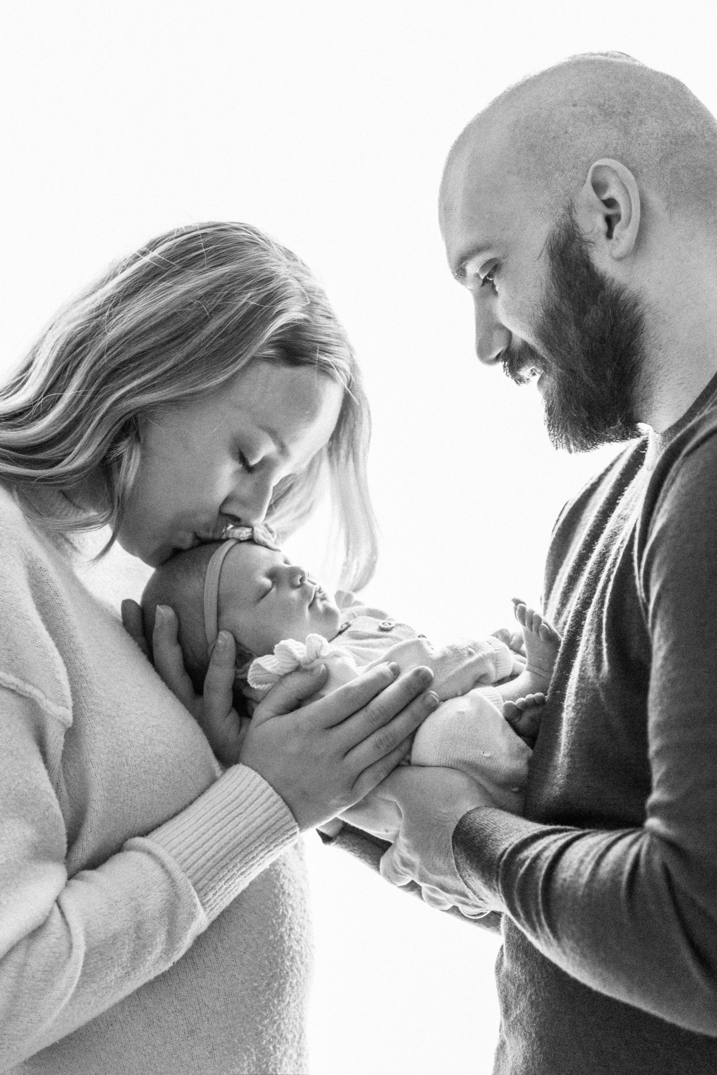 A black and white photo of a family holding a baby. The mother is kissing the baby's forehead, and the father is looking down at the baby, creating a tender moment.