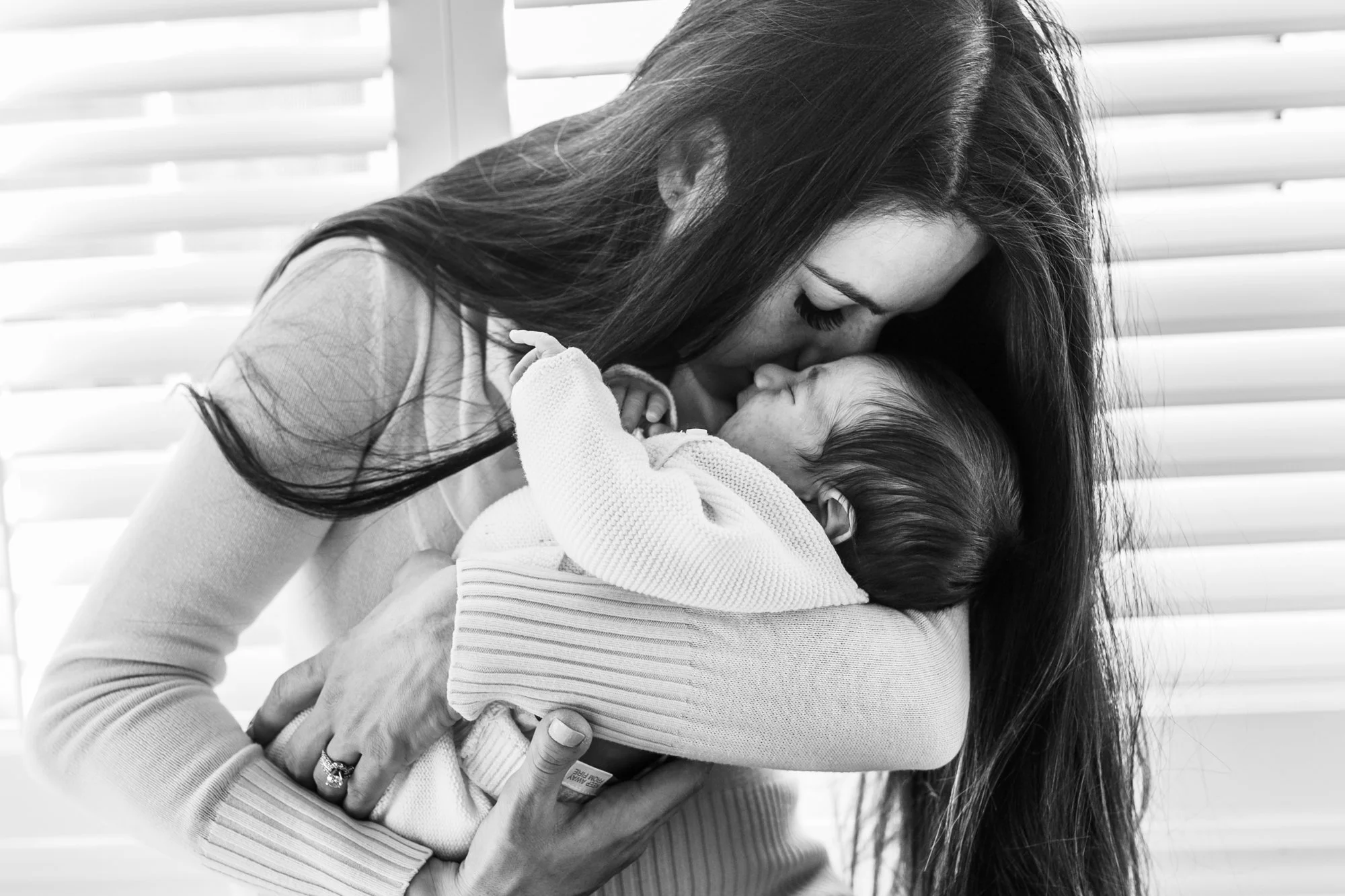 Black and white photo of a woman gently kissing a sleeping baby, holding the baby close to her chest, in front of a window with blinds.