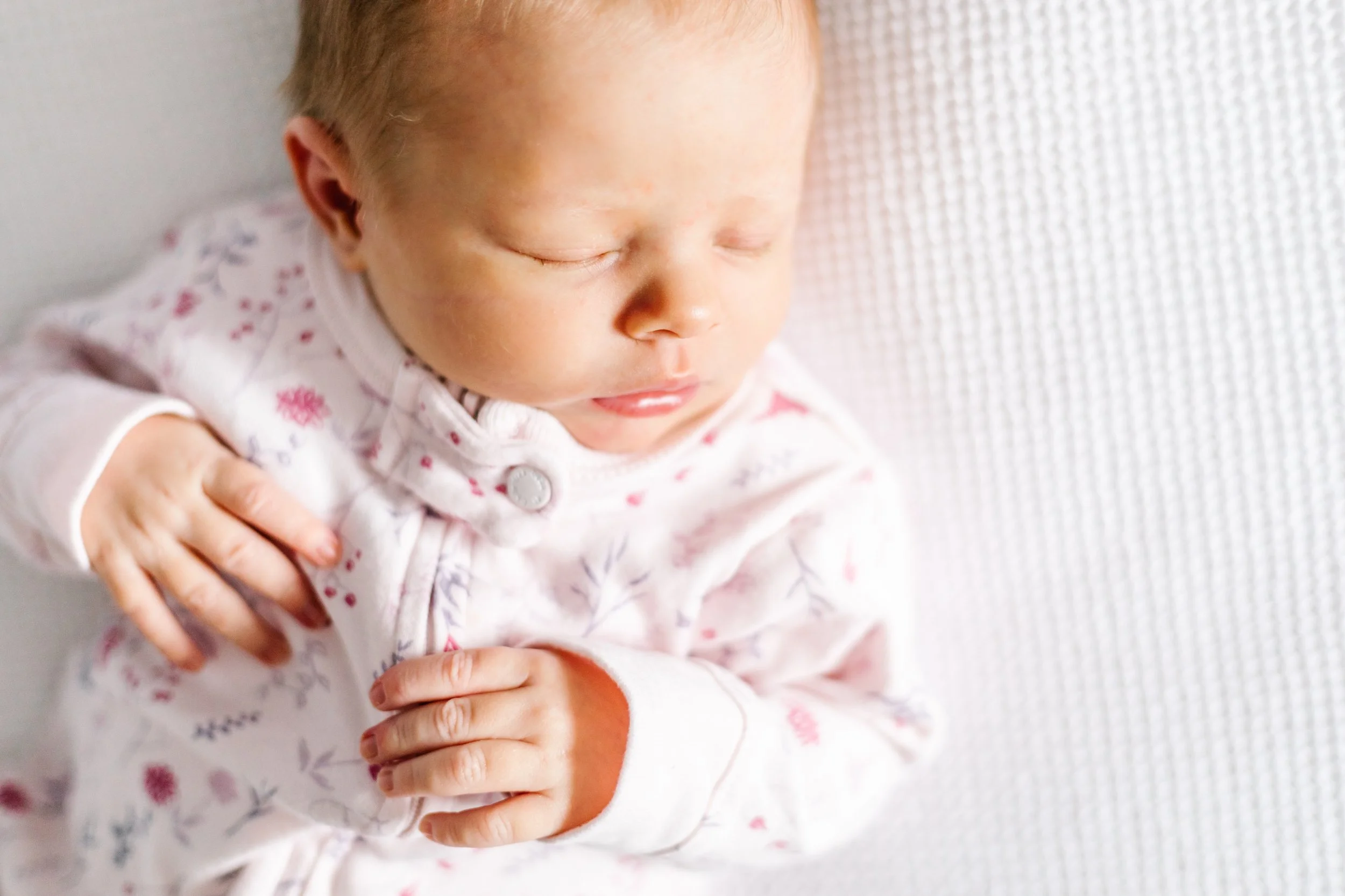 Sleeping newborn baby wearing a floral onesie on a white blanket.