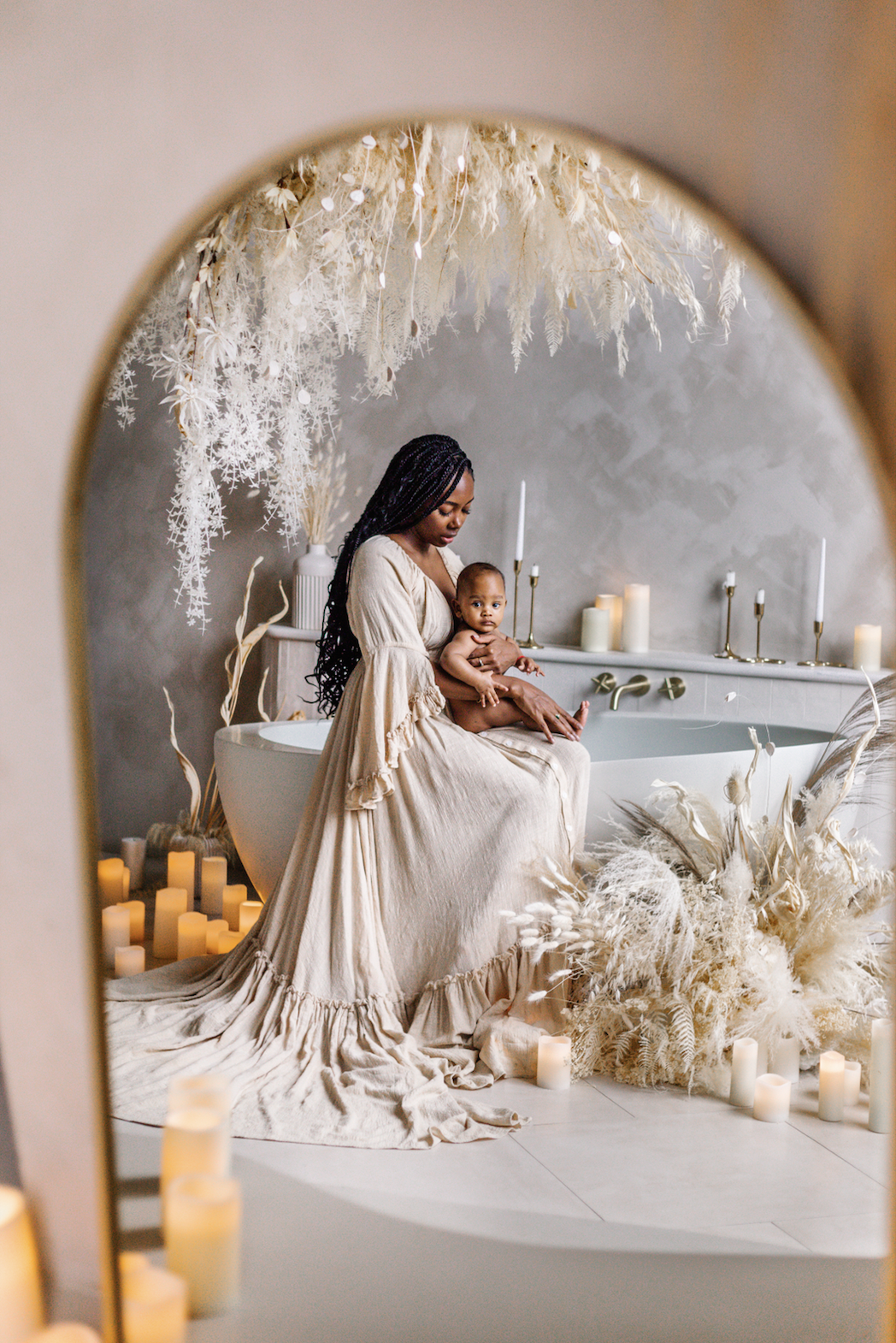 A woman wearing a long, flowing beige dress holds a baby while sitting on the edge of a bathtub. The scene is decorated with white flowers, candles, and pampas grass, creating a serene atmosphere.